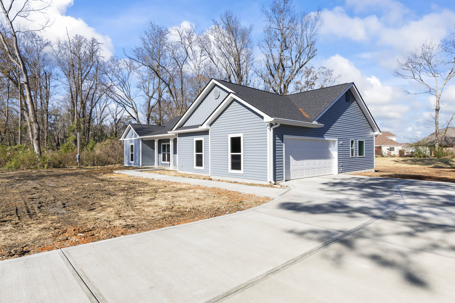 Concrete driveway bordered by green lawn leading to a modern home with white siding, large windows, and attached garage; mature trees in the background under partly cloudy sky.