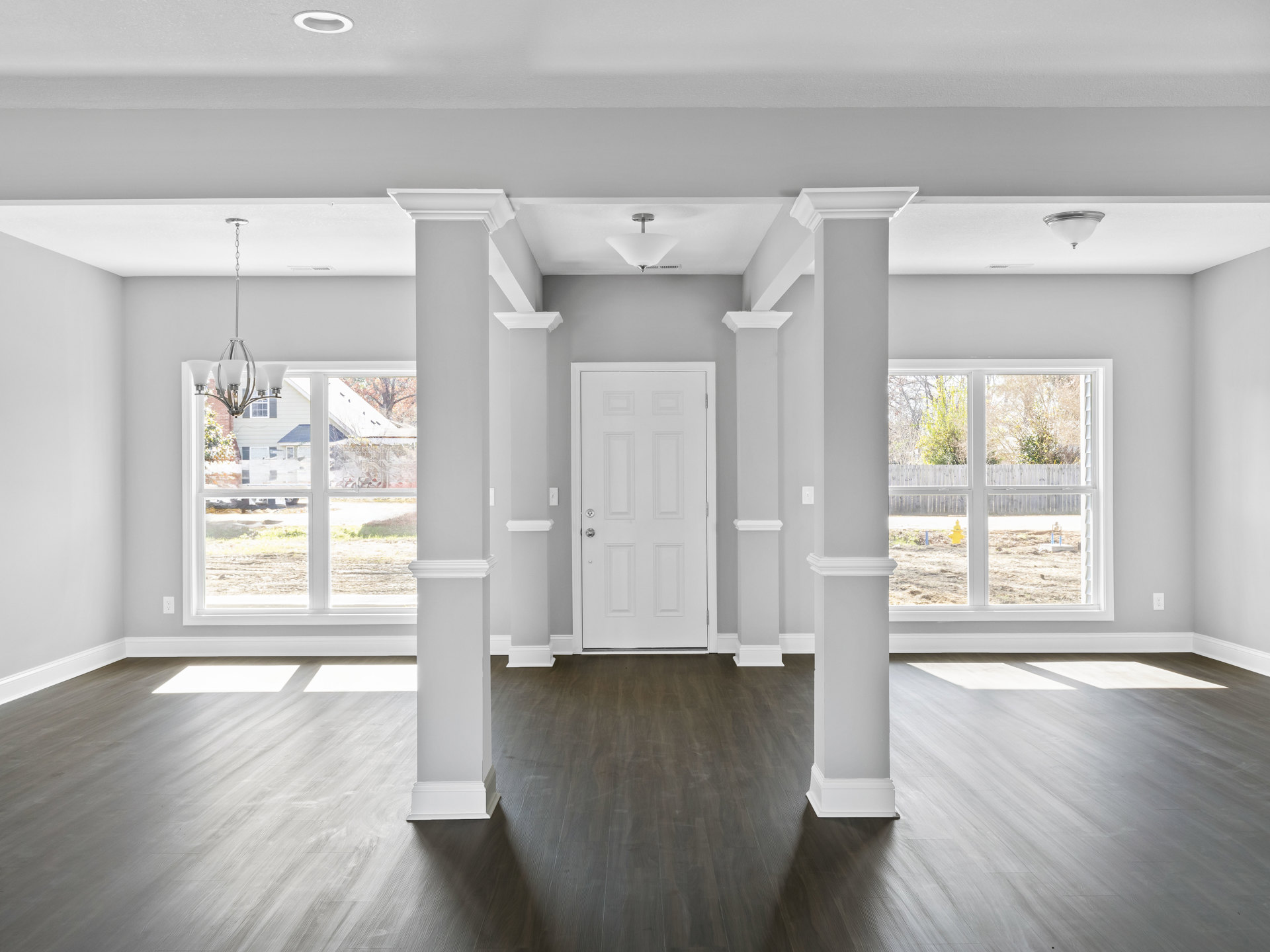 Dark wood flooring, white columns, white walls, white door with silver doorknob, chandelier, window, ceiling molding