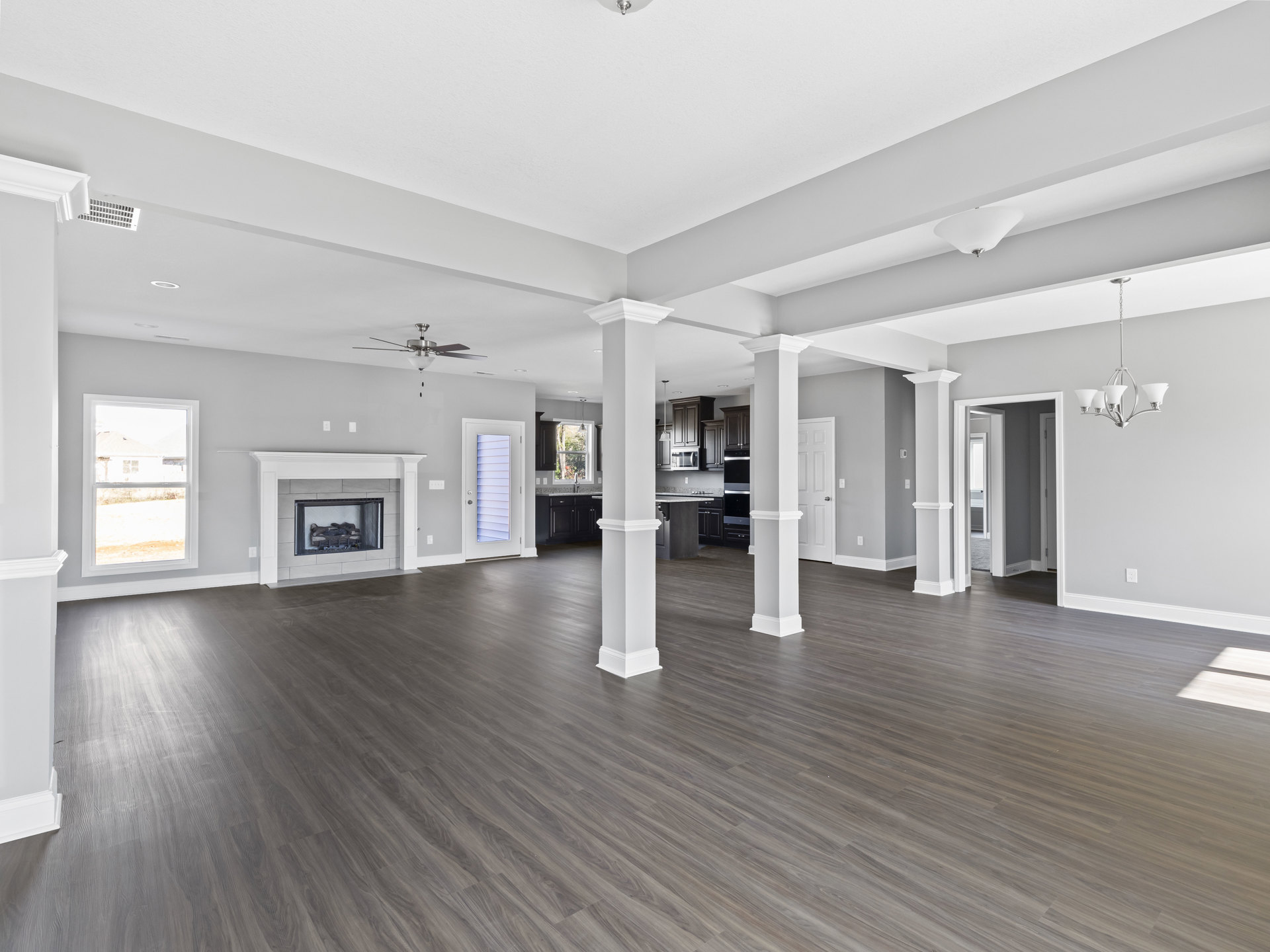 Spacious living area with white columns, wood flooring, glass fireplace door, large window overlooking neighboring house, and adjacent kitchen with white walls and pillar.
