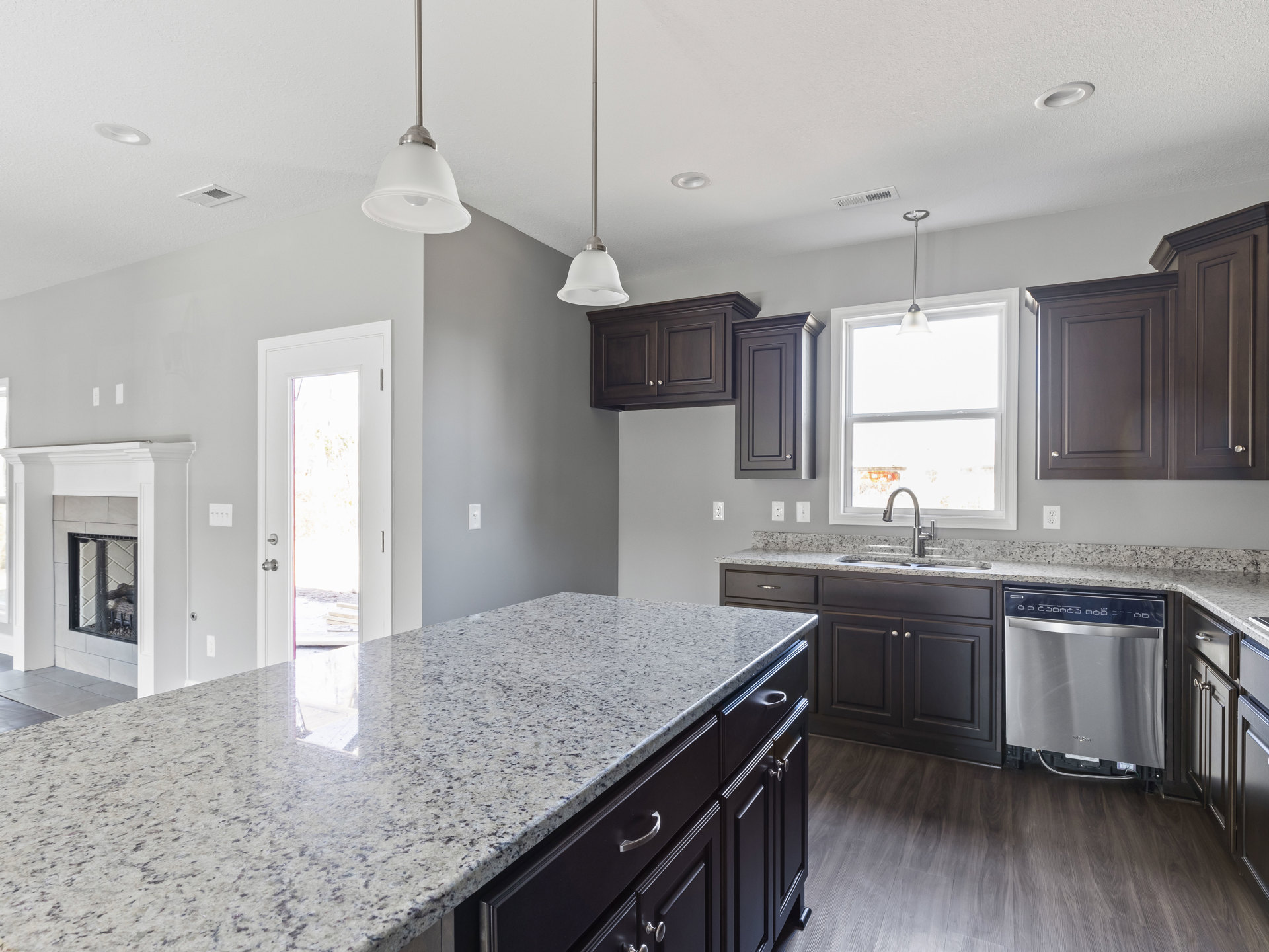 Granite countertops, dark wood cabinets, stainless steel dishwasher, open cabinet door, white door with window, globe light fixture, sunlight streaming through window