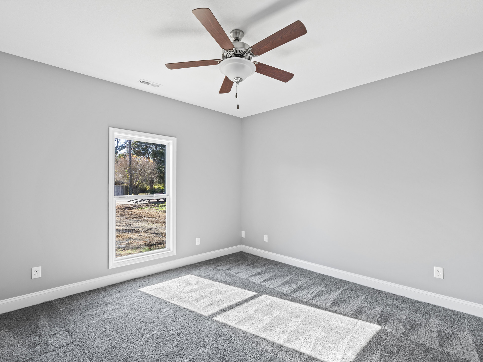 Ceiling fan with light fixture above grey carpet featuring white squares, window overlooking dirt yard, white plaster walls