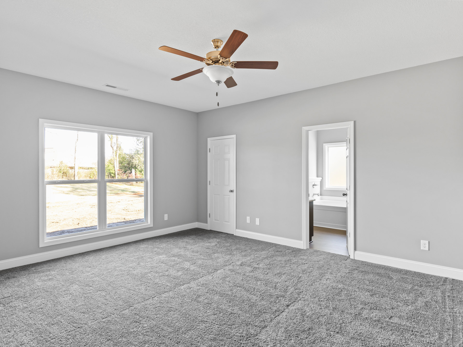 Carpeted room featuring a ceiling fan with light fixture, white walls, large windows overlooking trees, and a white door with silver knob; adjacent bathroom visible with white