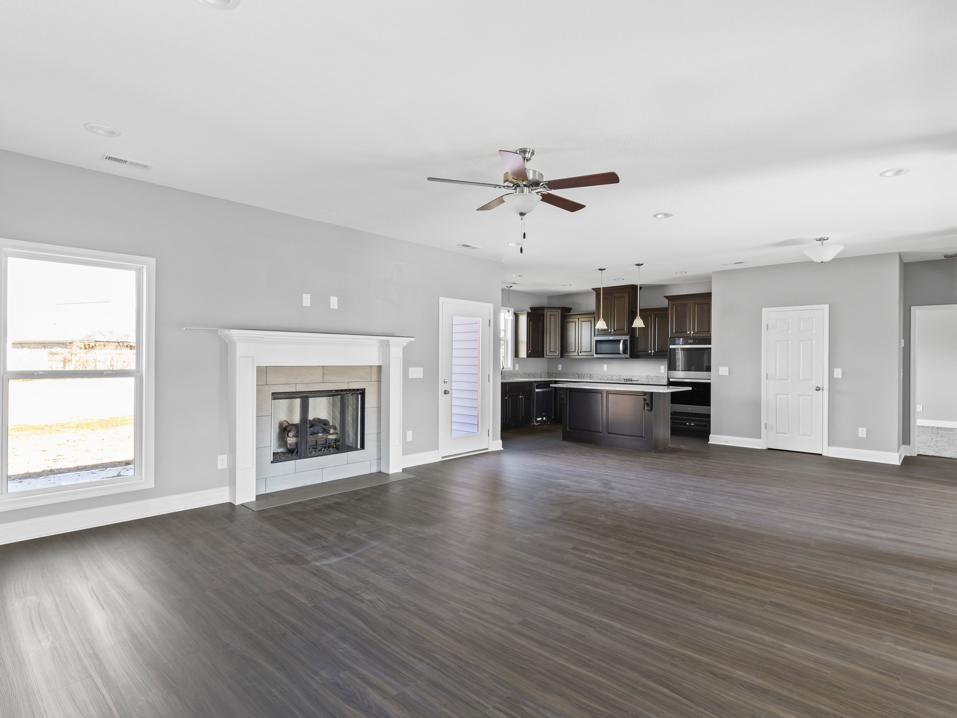 Spacious living room featuring hardwood floors, central fireplace with glass doors, ceiling fan with light fixture, large window showing neighboring house, and white door with