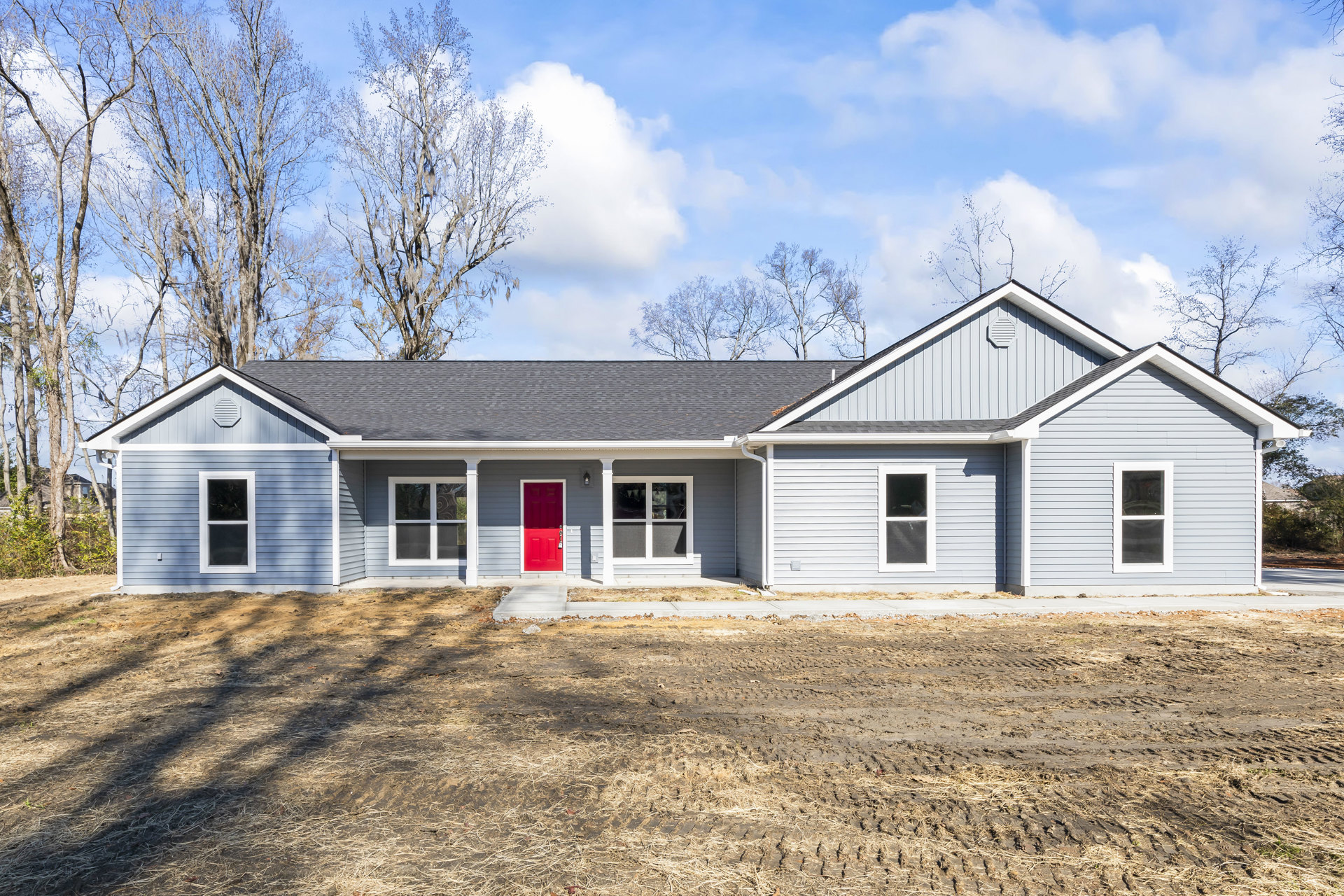Two-story cottage with white siding, red front door, white-framed windows, black window screens, surrounded by trees, set along a dirt road under a cloudy sky
