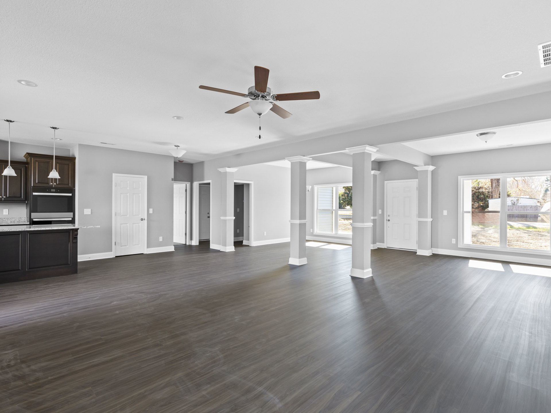 Spacious room with wood flooring, white columns, ceiling fan with light fixture, white doors with silver handles, and a window letting in natural light