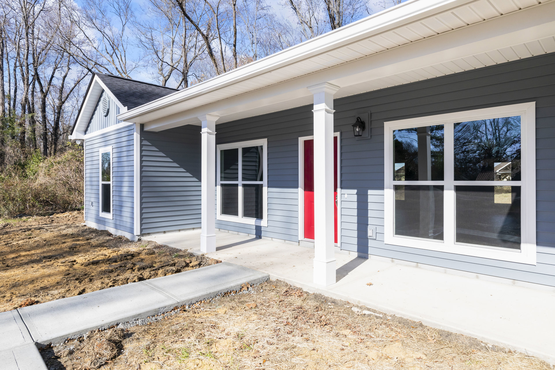 White siding house with red front door, white pillars supporting porch roof, window with closed blinds, concrete walkway bordered by grass and dirt, tree visible in background.
