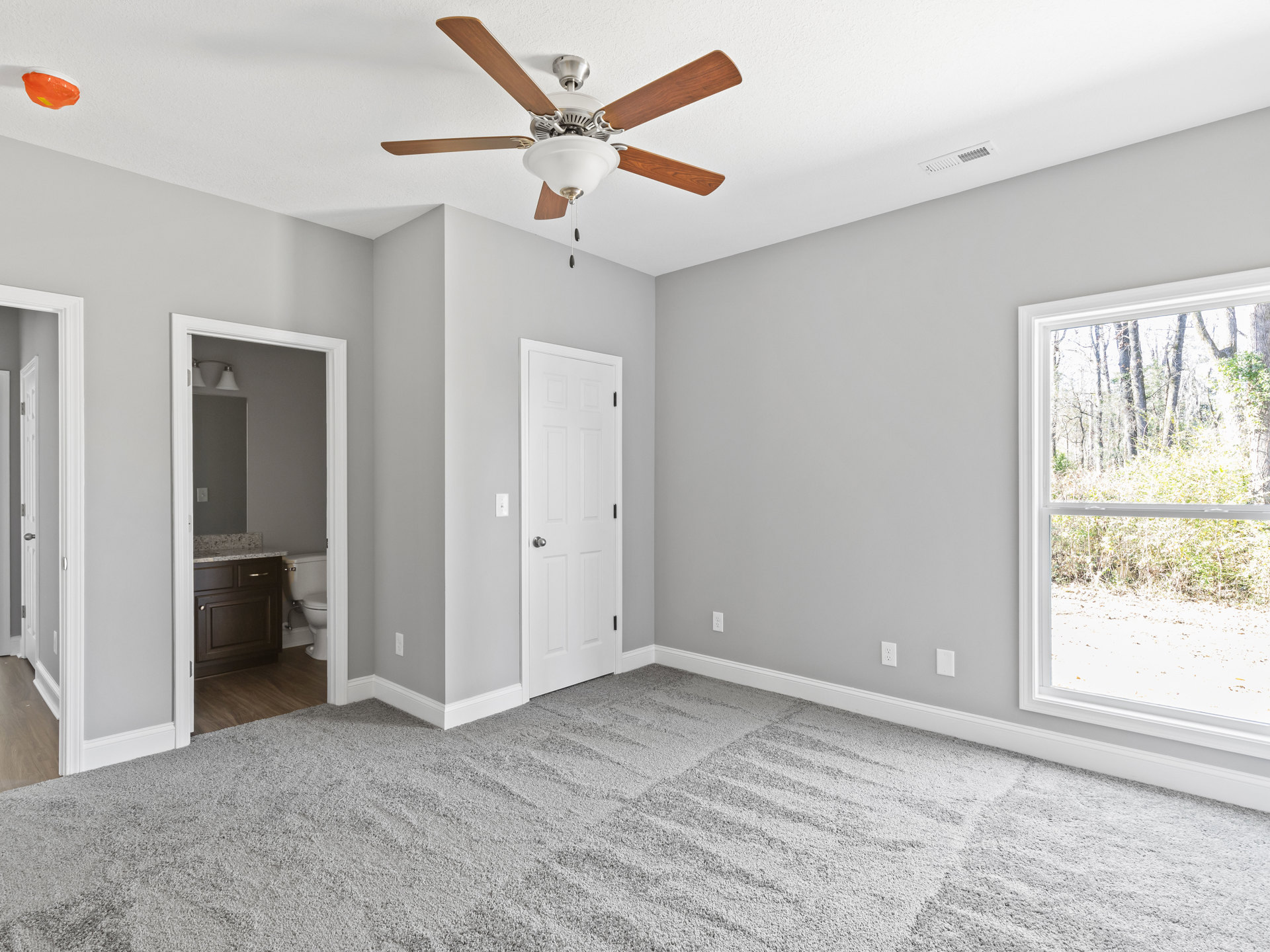 Carpeted bedroom with ceiling fan and light fixture, white door with silver knob, window overlooking trees, adjacent bathroom with sink and toilet
