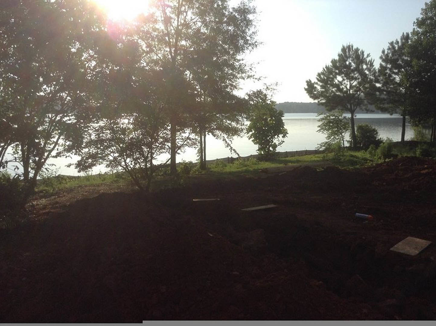 Dirt field bordered by green trees, sunlight filtering through leaves, lake visible in the background, clear morning sky overhead