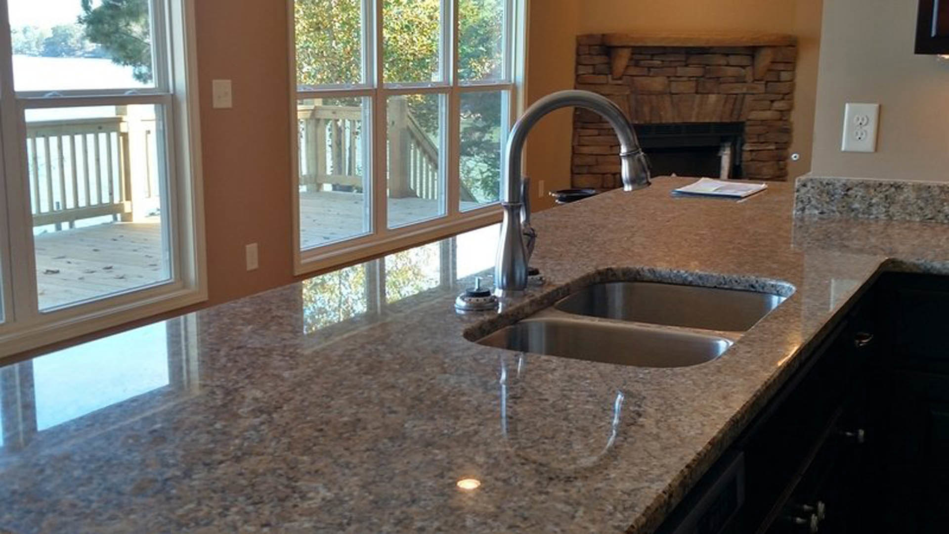 Granite kitchen counter with double stainless steel sink, tile backsplash, built-in outlet, and fireplace in the background