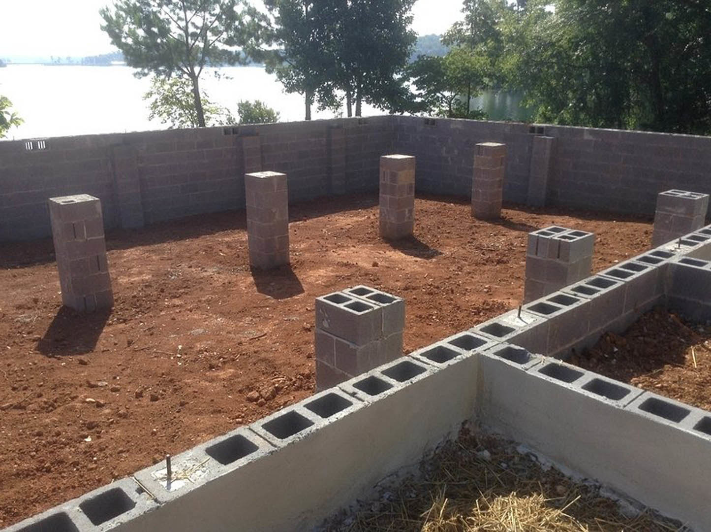 Concrete block foundation wall with open holes, stacked grey bricks, brick pillar, dirt ground, trees and sky in background