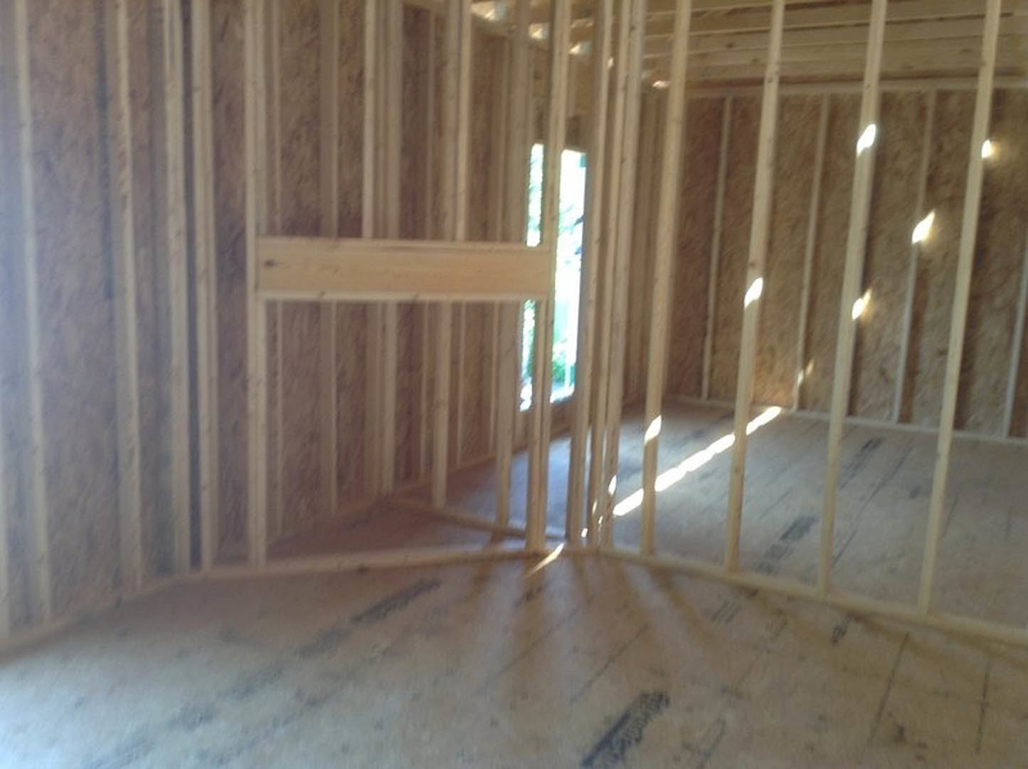 Room under construction with exposed wood framing, plywood ceiling, hardwood plank flooring, and visible wood beams; light illuminates surfaces, with a blurry figure partially