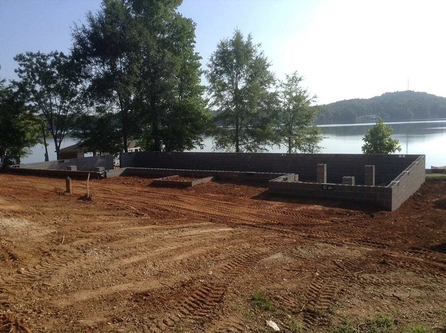 Concrete foundation wall on dirt construction site near pond, surrounded by green trees and grassy landscape