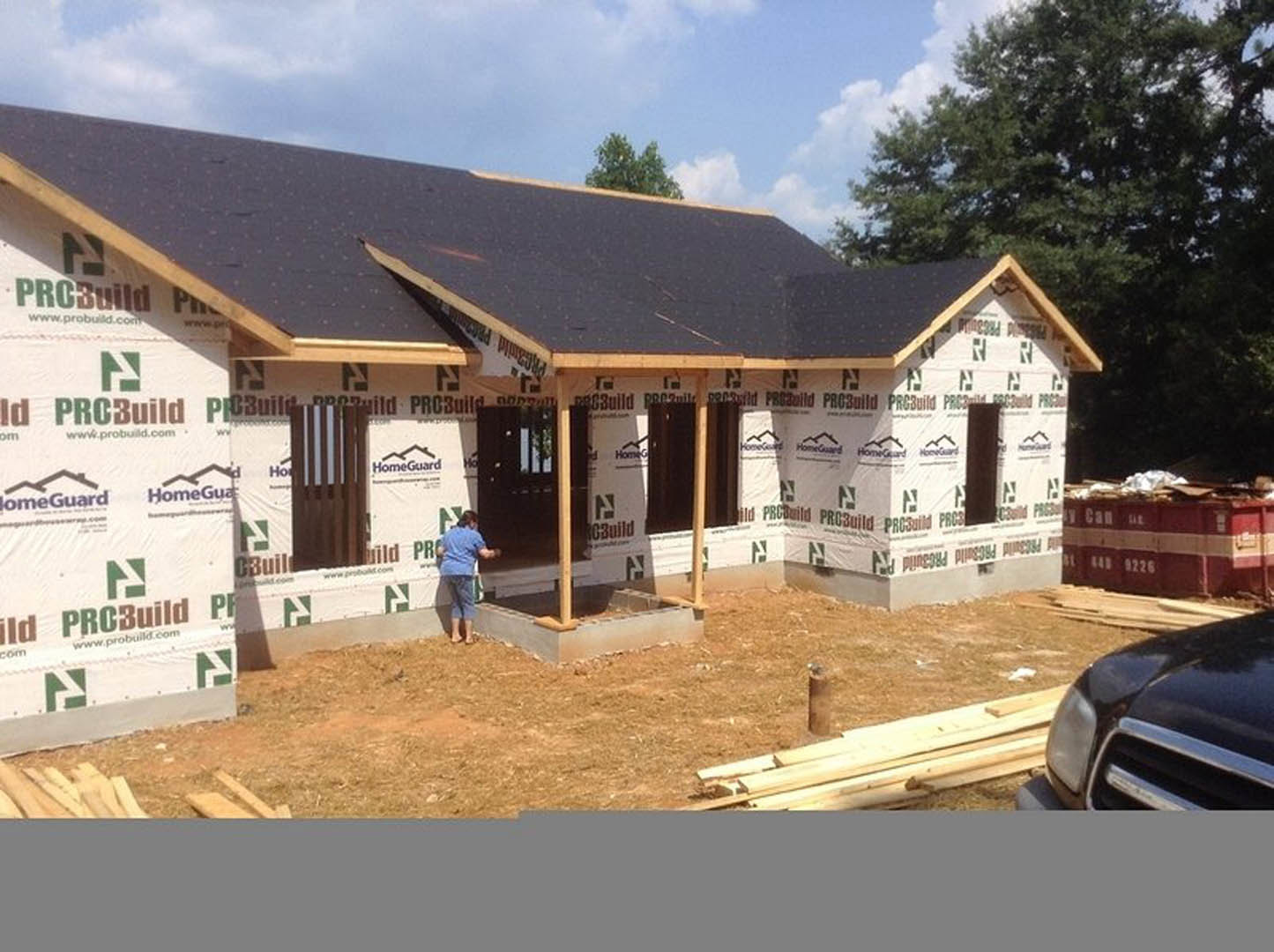 Person in blue shirt standing near partially built house with exposed framing, construction materials, parked car, red and white container, and green logo sign on exterior