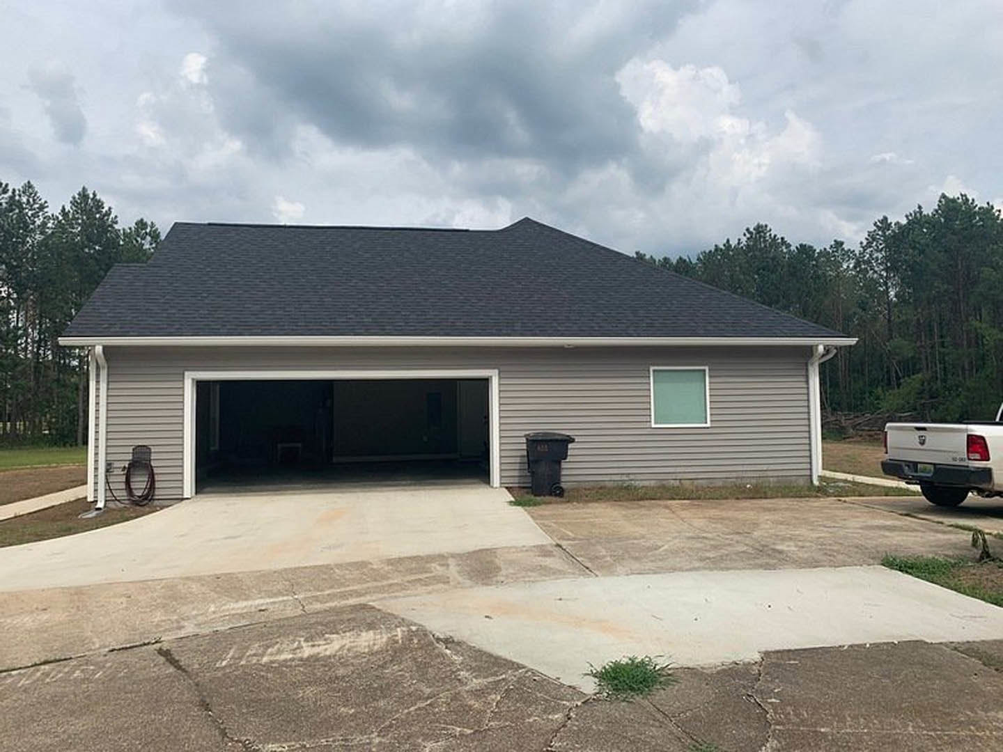 Two-story custom home with attached garage, white siding, gray roof, driveway with parked white truck, black garbage can near garage, green bush in dirt yard, large windows, cloudy