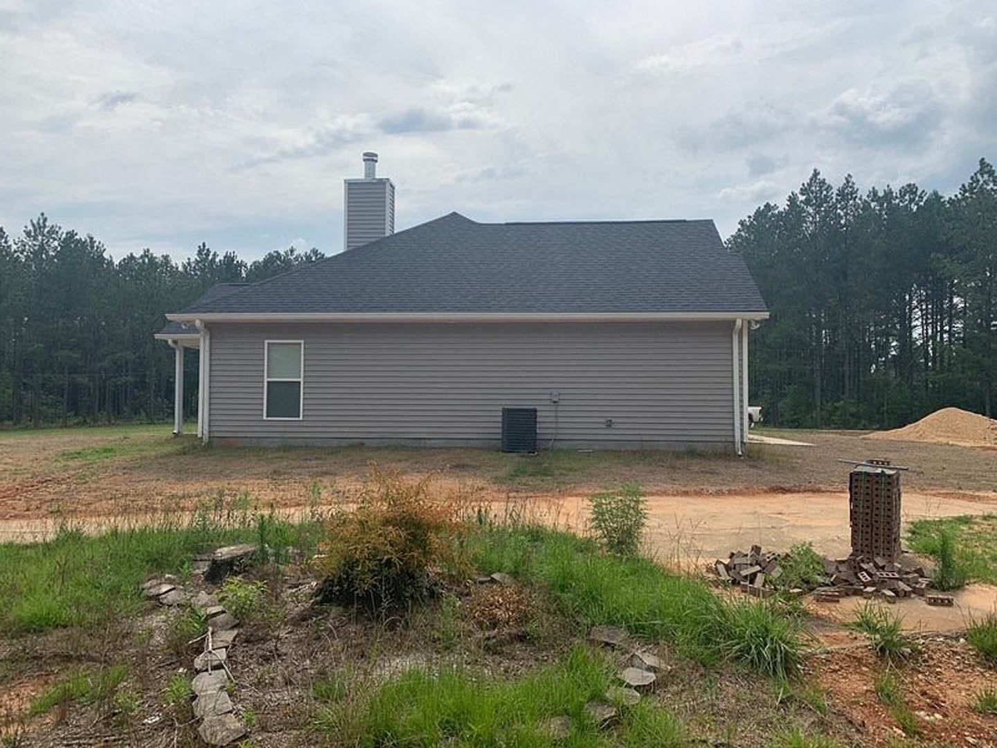 Two-story cottage with white-framed windows, gray siding, and brick chimney at the rear, surrounded by grass and trees under a cloudy sky