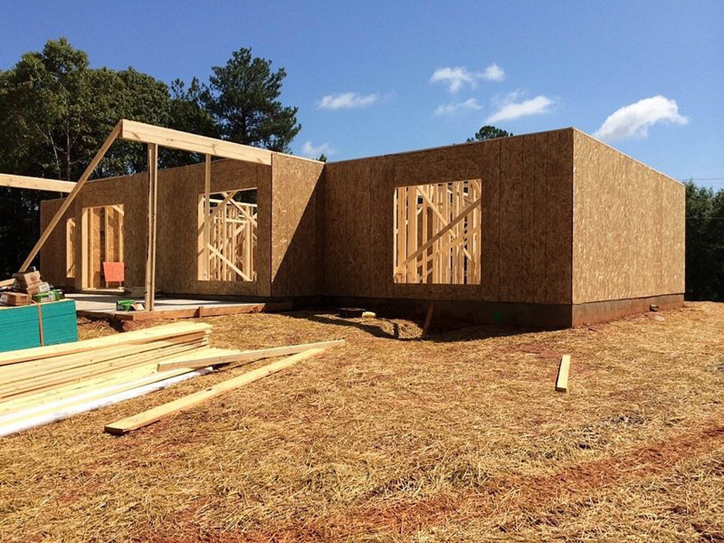 Wood-framed house under construction with exposed beams, dry grass and lumber scattered on the ground, trees and cloudy sky in the background