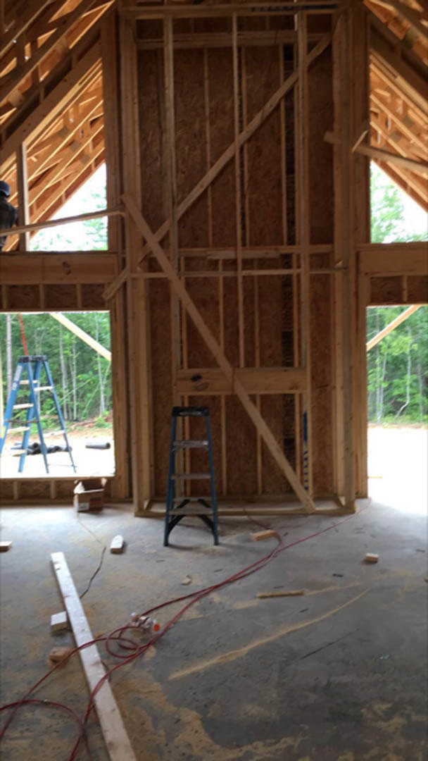 Wood-framed house under construction with exposed beams, blue ladder, red electrical cable on the floor, and worker in helmet assembling structure