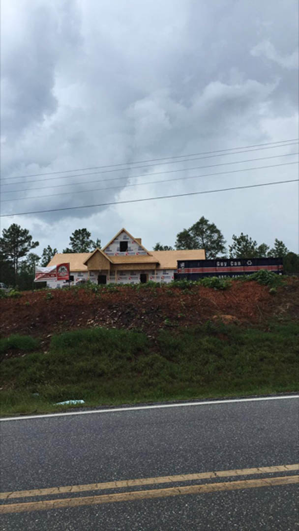 Framed house under construction on a dirt hill, surrounded by trees and grass, cloudy sky overhead, power lines visible