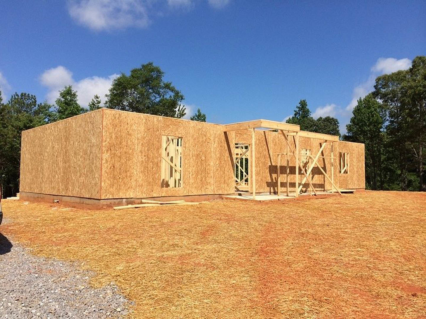 Wood-framed house under construction on grassy lot, surrounded by trees, with blue sky and clouds overhead