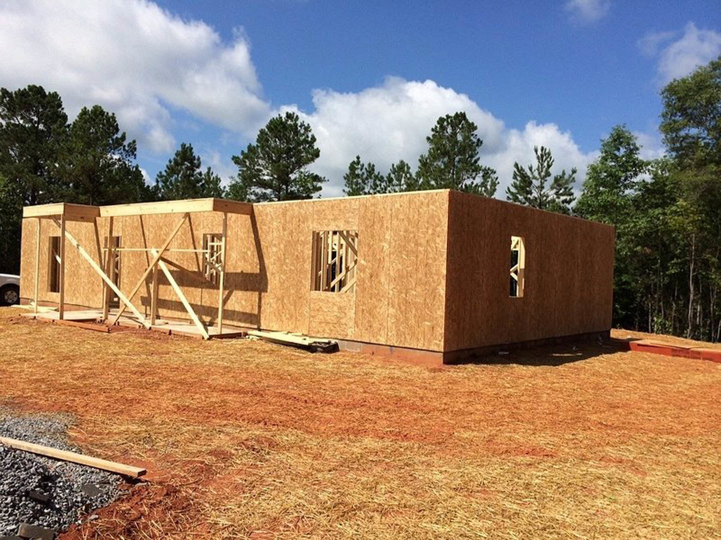 Wood-framed house under construction with exposed beams, unfinished window opening, gravel foundation, grassy lot, and trees in the background under a partly cloudy sky
