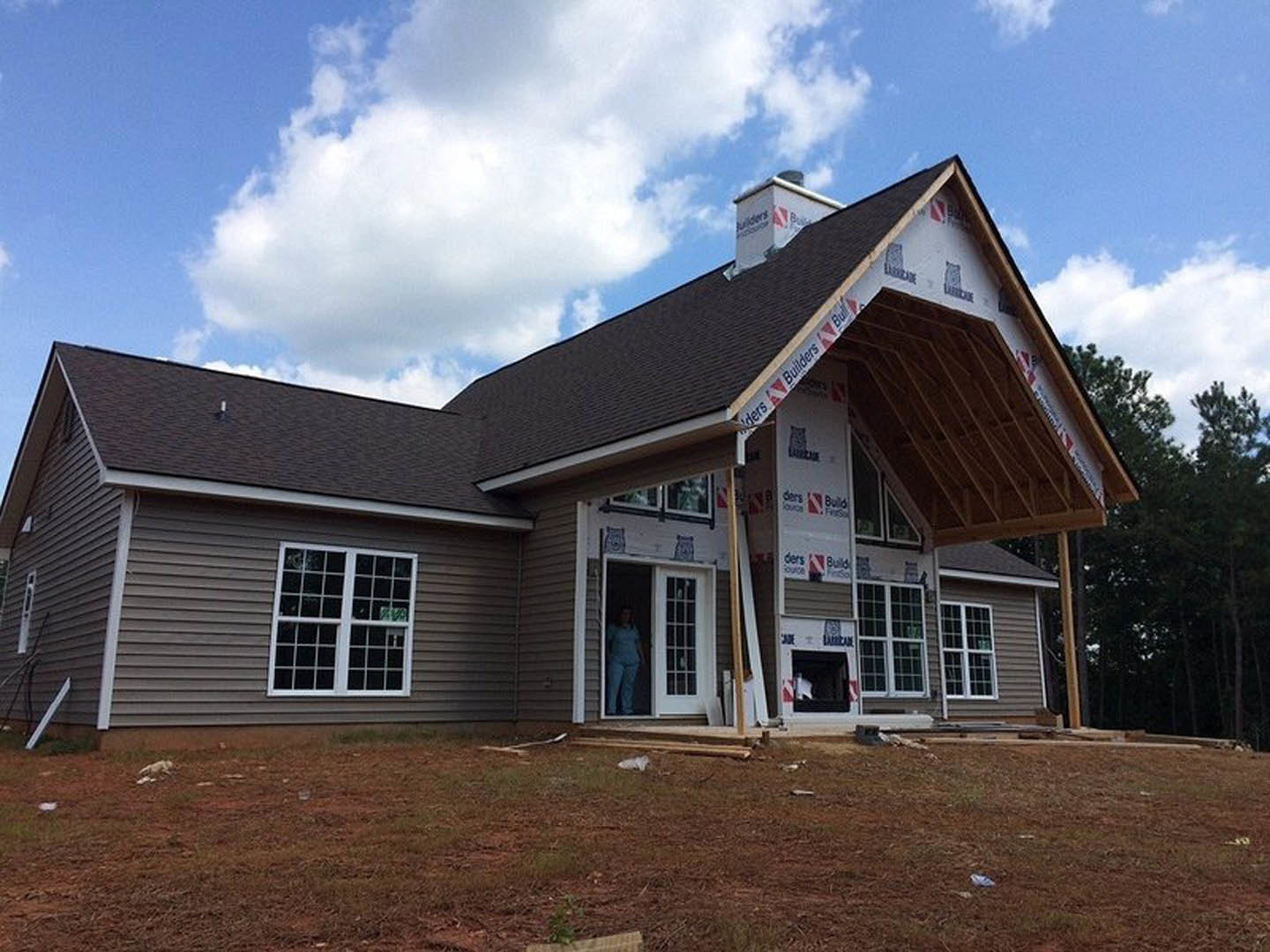 Framed house under construction with exposed wood, multi-pane window, dirt patch scattered with debris, man in blue pants standing in doorway, white metal bar visible on roof