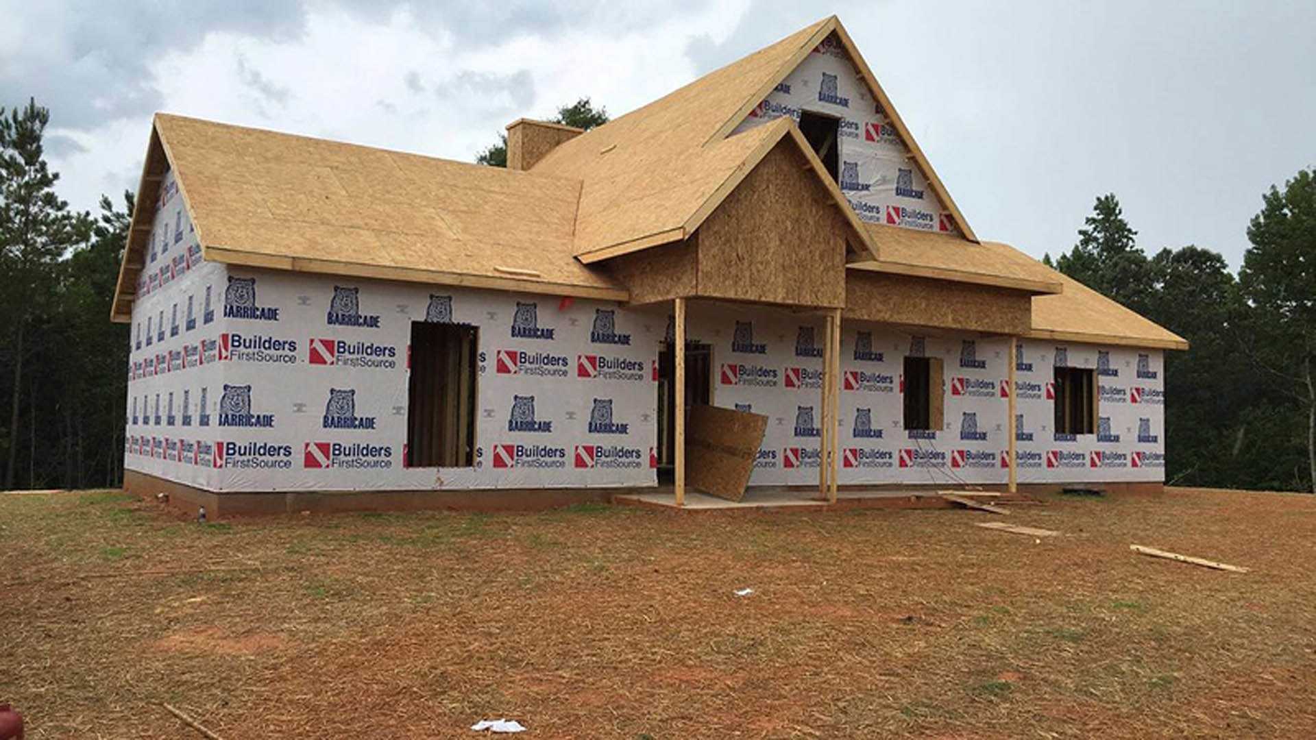 Partially built house with wood panel siding, several installed windows, and a black bear logo displayed on a white wall; surrounded by trees under a cloudy sky.