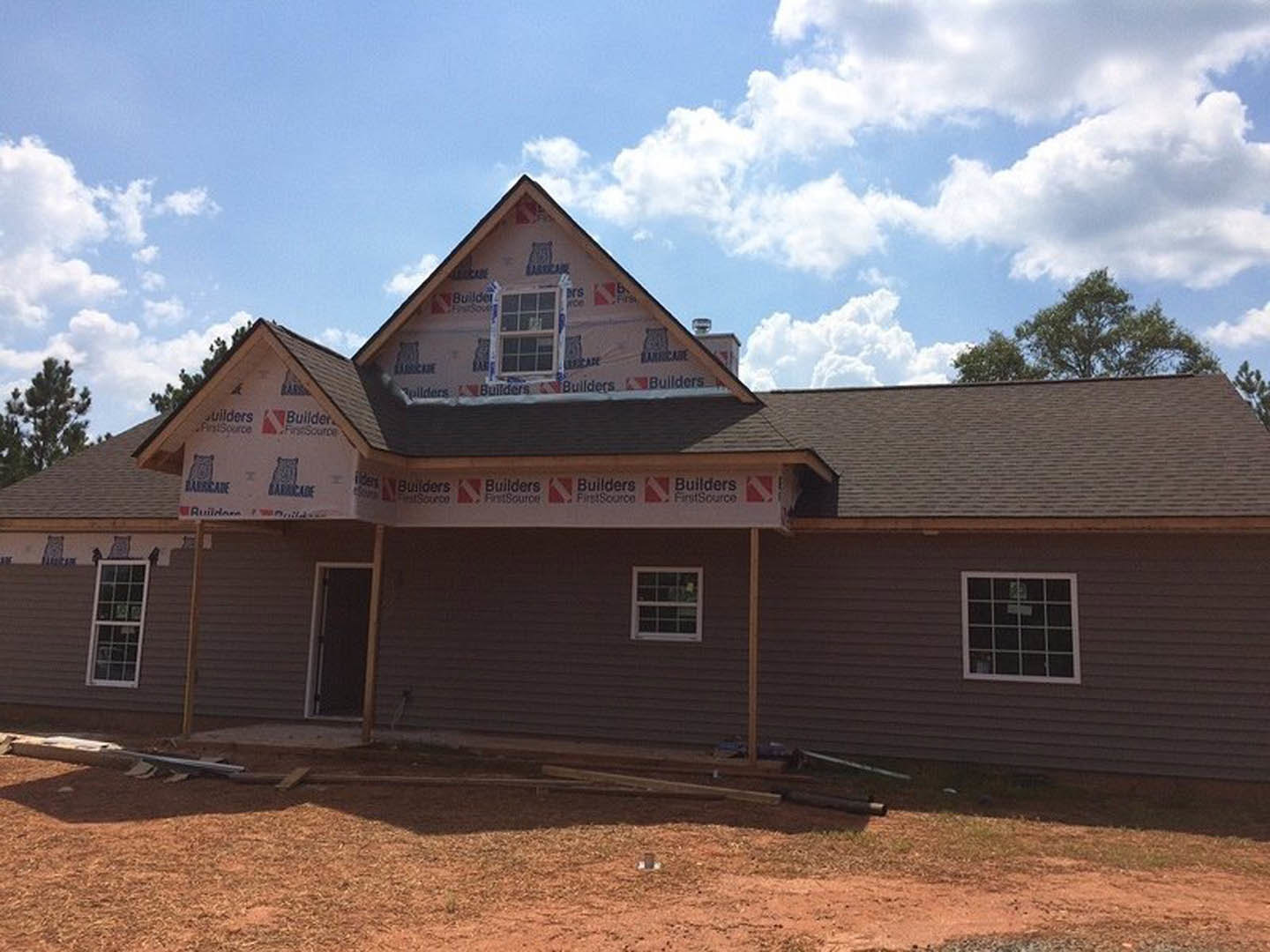 Two-story house under construction with gray shingle roof, white-framed multi-pane windows, dark entry door with white trim, triangular dormer window, surrounded by trees and