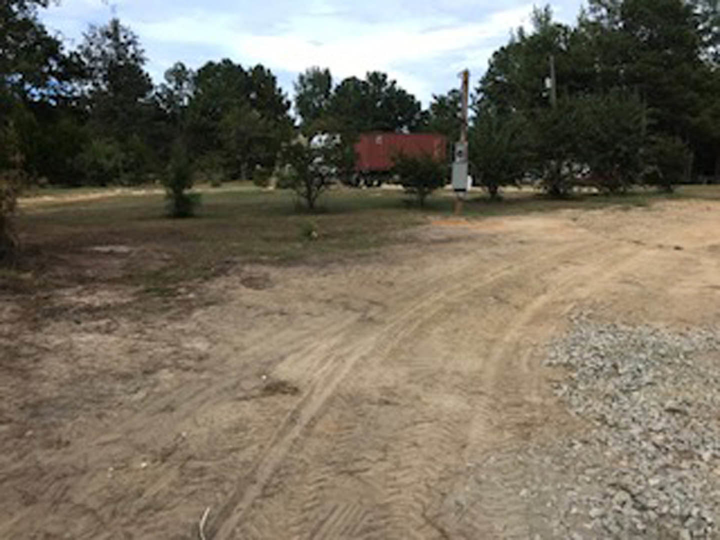 Dirt road with tire tracks leading through a wooded area, red trailer and dense trees in the background, cloudy sky overhead