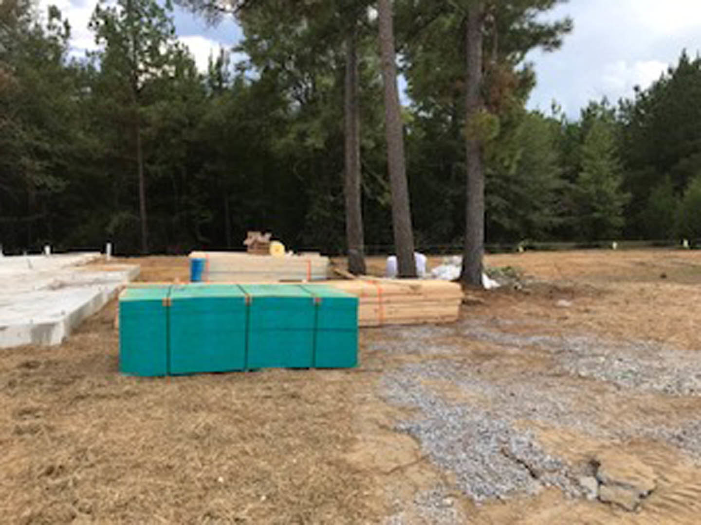 Stacked lumber and concrete blocks on bare ground near trees under open sky