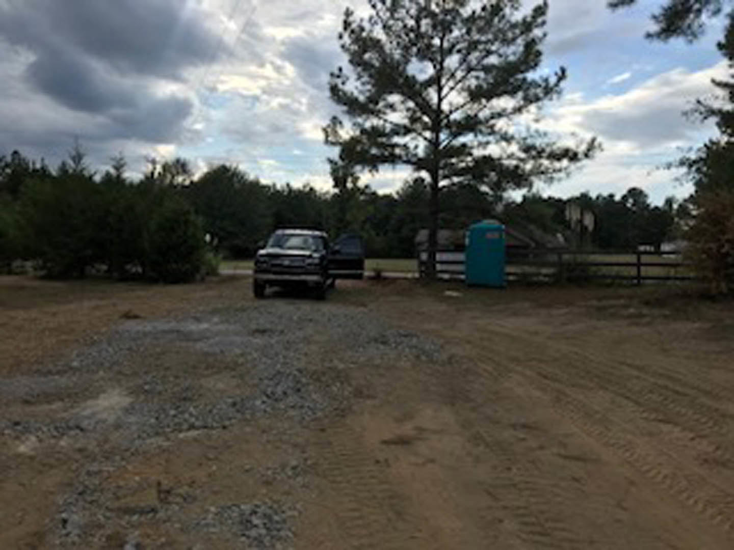 Pickup truck parked on dirt lot beside grassy area with mature tree under partly cloudy sky