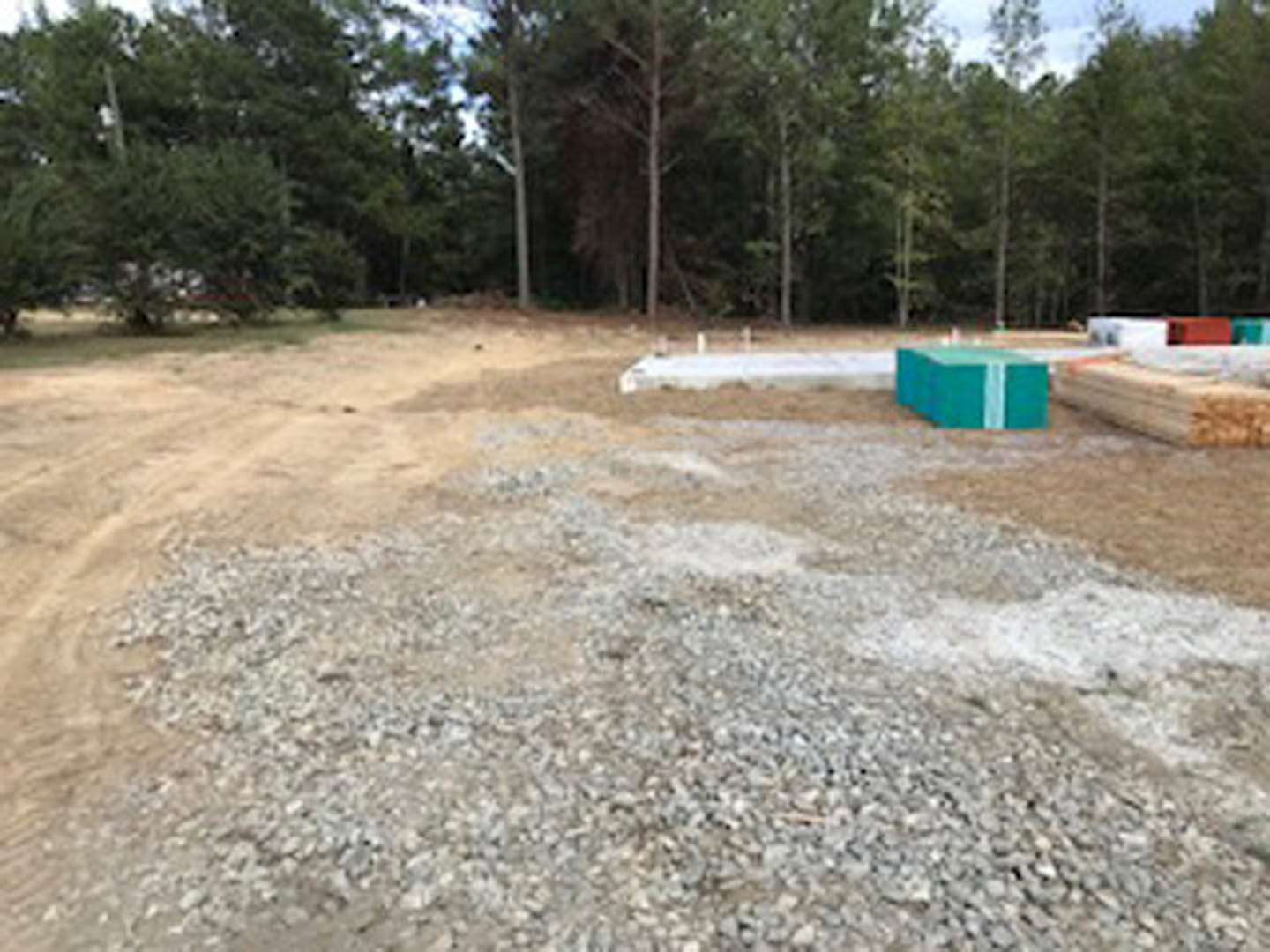 Gravel and dirt clearing with a blue utility box, surrounded by mature trees under a clear sky