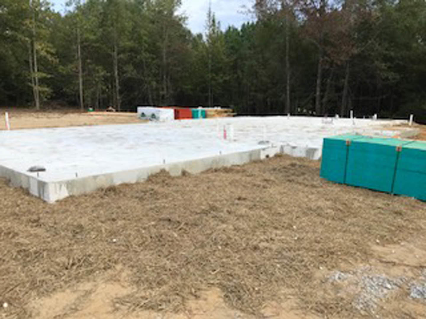 Concrete foundation with central blue utility box, surrounded by soil and grass, trees in background, clear sky overhead