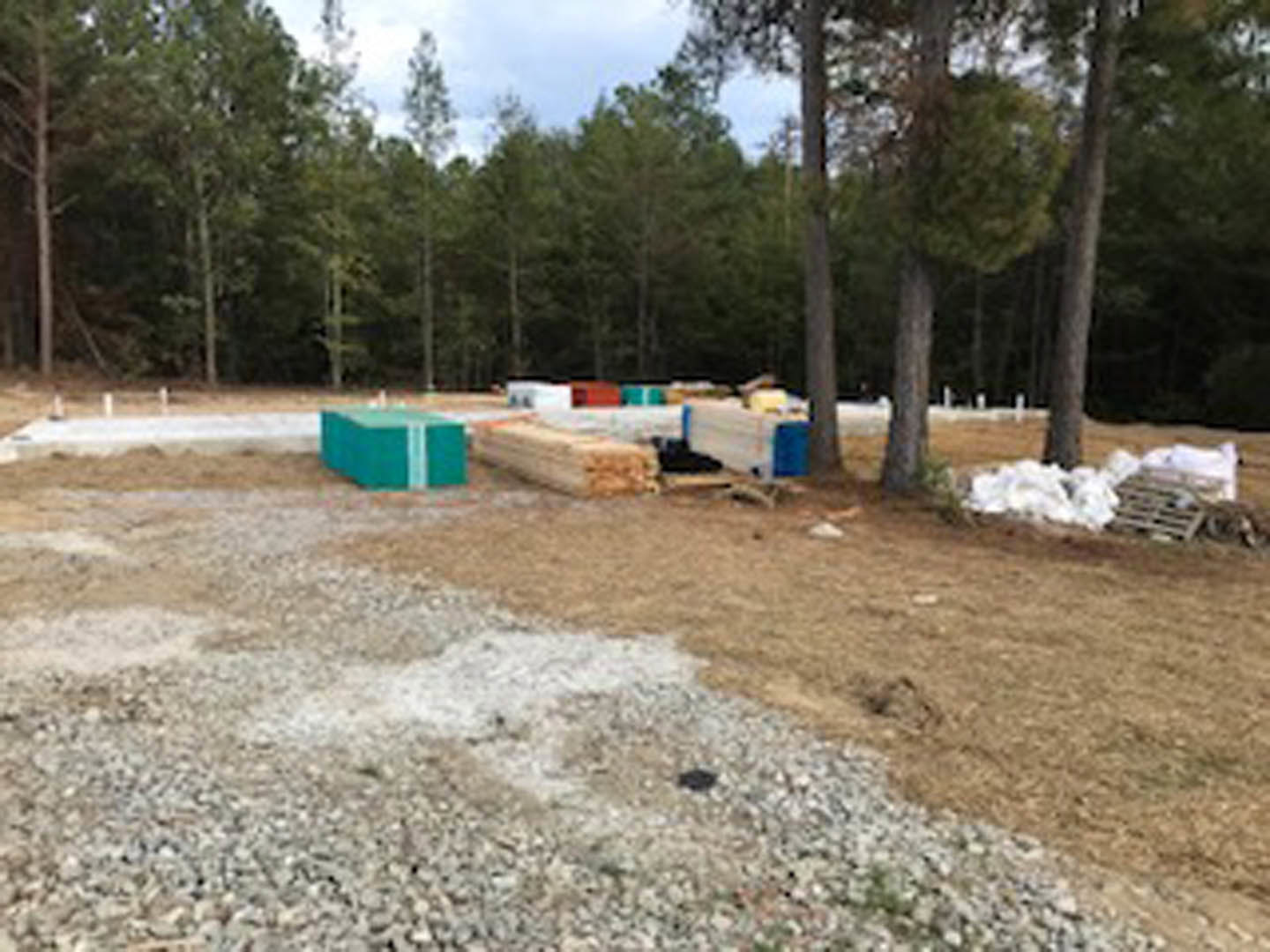 Stacked lumber, building supplies, and rocks on dirt and gravel in an open field bordered by trees and grass under a pale sky