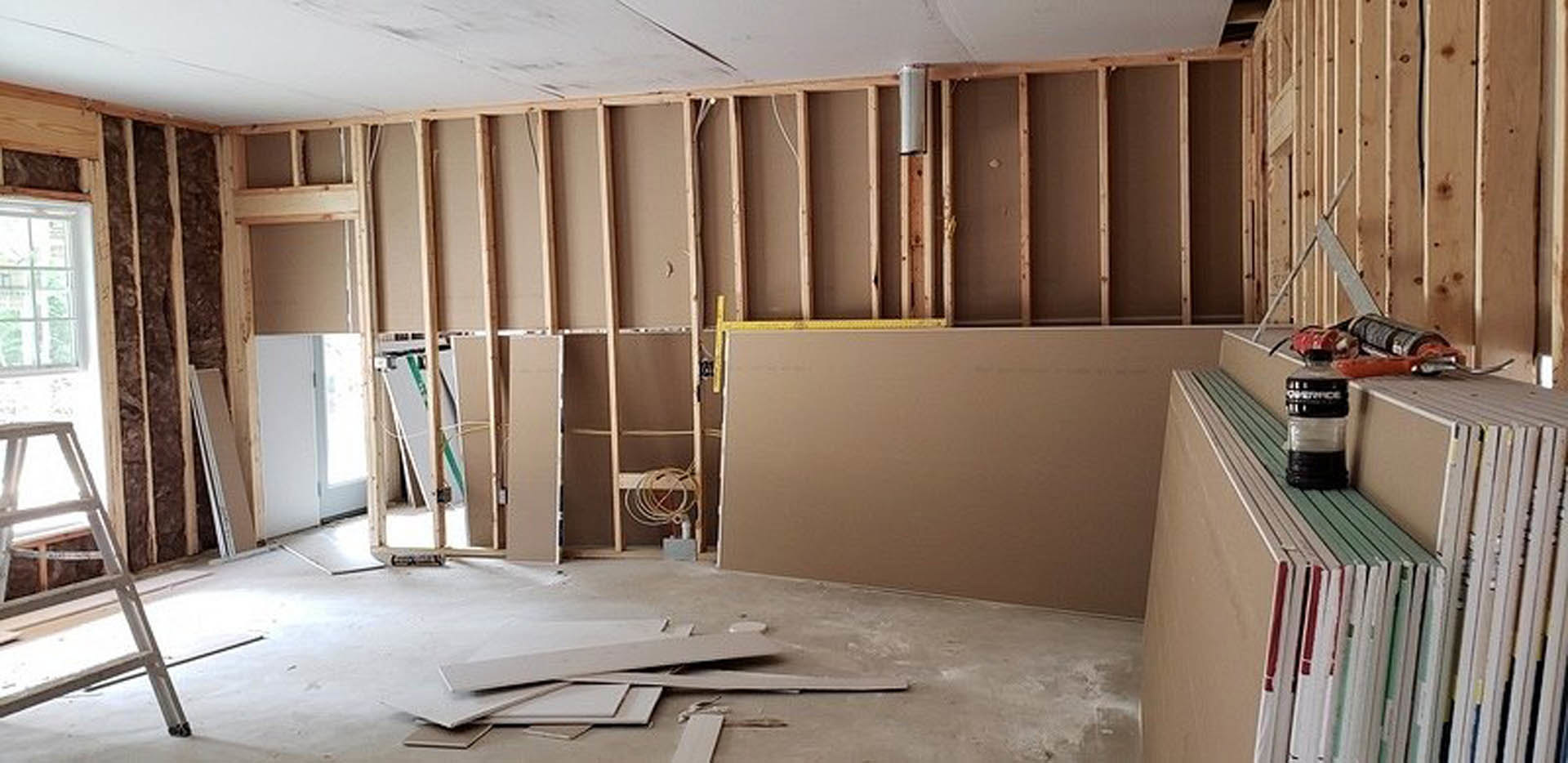 Wood-framed room under construction with exposed beams, yellow measuring tape on the floor, ladder leaning against a window, close-up of unfinished wall, bottle on a surface, stack