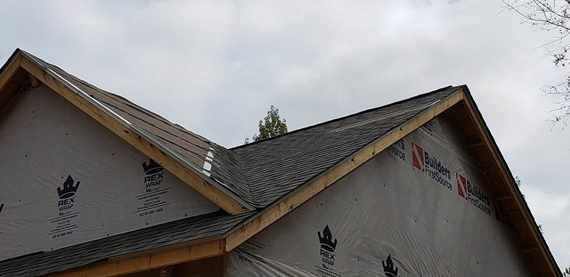 Partially finished roof with exposed wooden framing and shingles, exterior walls with white siding, windows installed, cloudy sky overhead