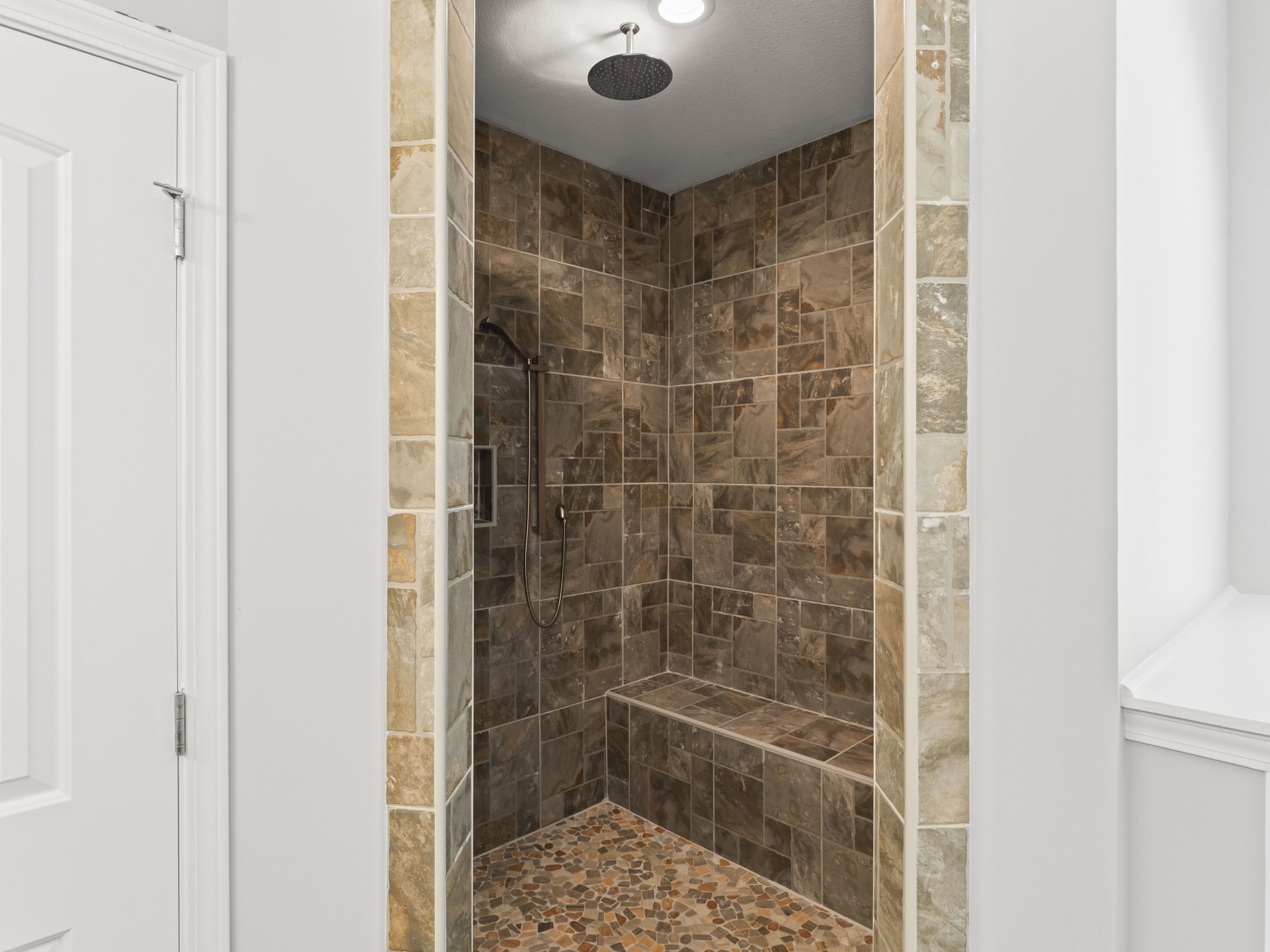 Walk-in shower featuring a textured stone wall, ceiling-mounted shower head, tile flooring, and a white built-in shelf adjacent to a white door with metal hardware.