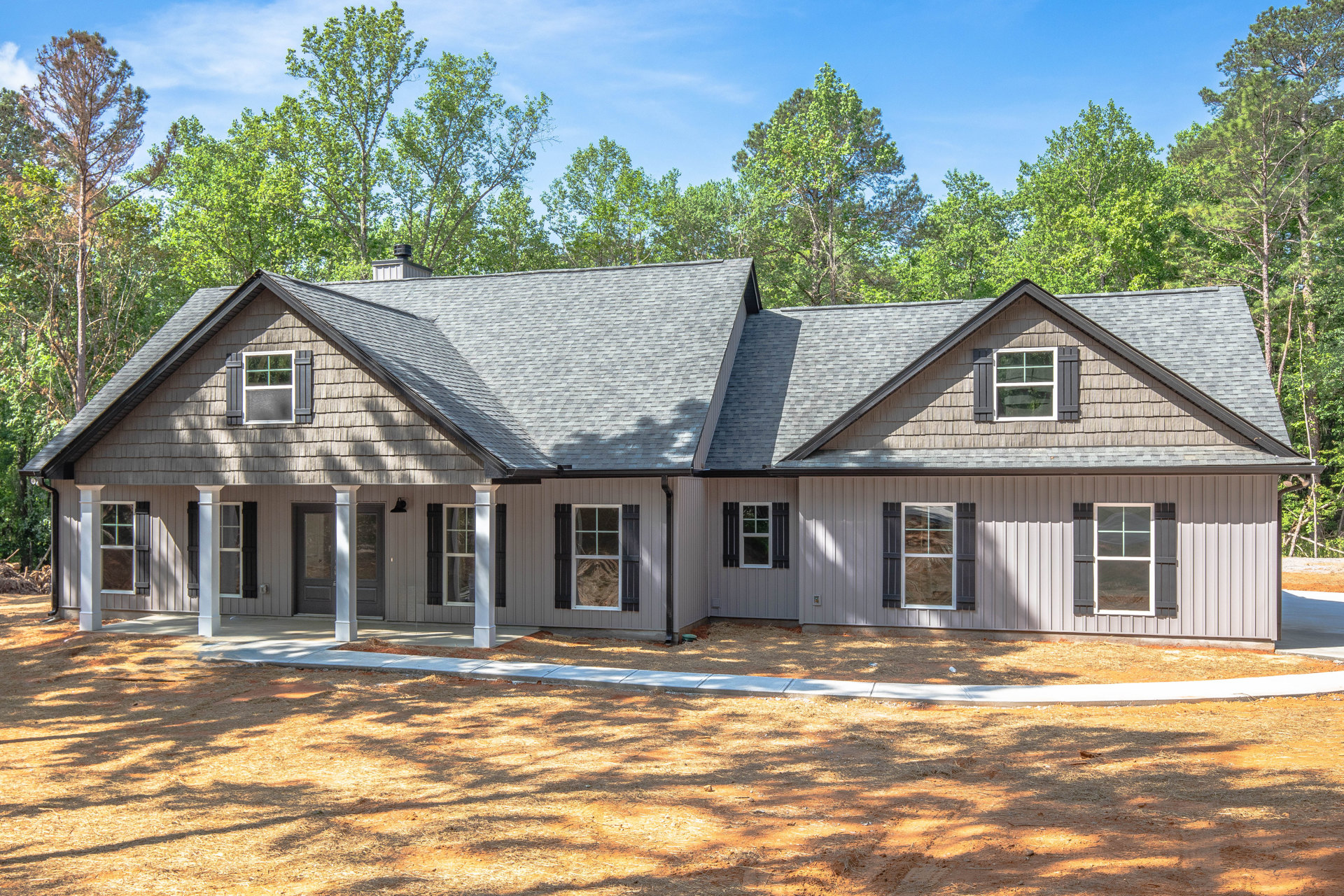 Two-story house with white columns, gray roof, large windows reflecting trees and sky, paved driveway, mature trees in background