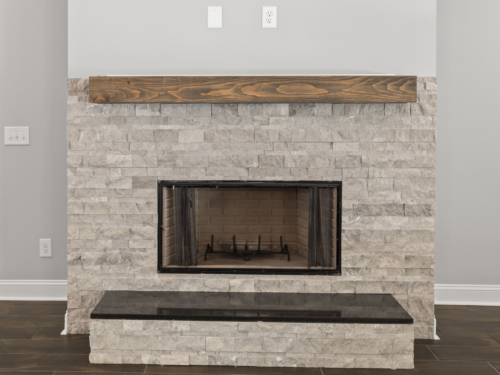 Stone fireplace with wood mantle, black fire screen, metal log rack, white rectangular light switch, and black ledge on hearth.