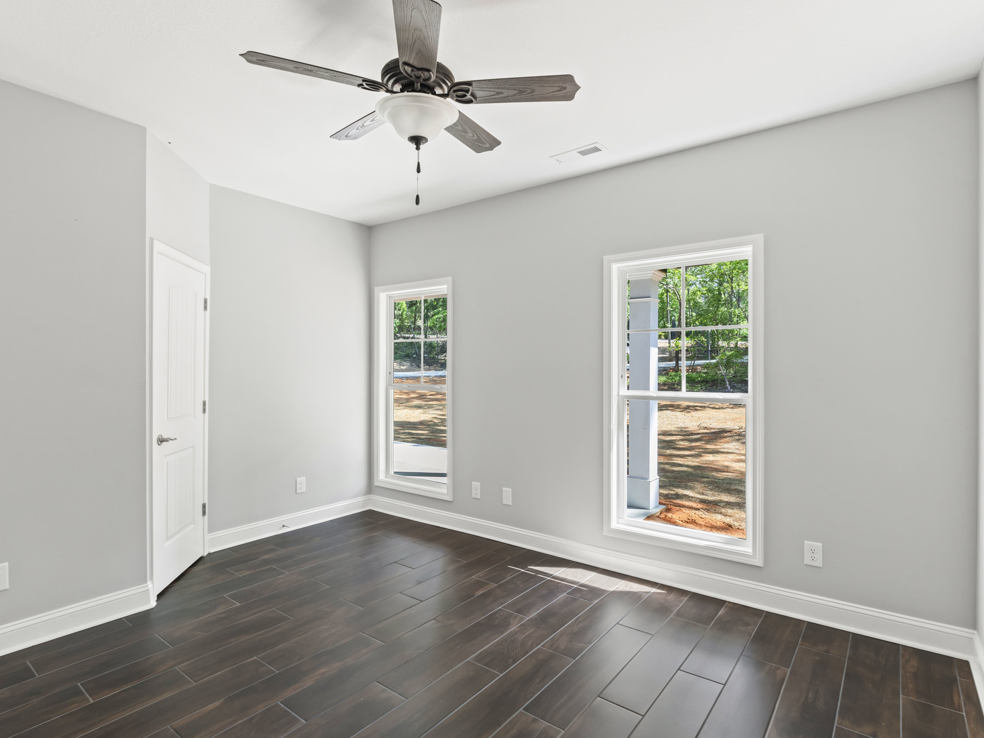 Ceiling fan with light fixture, wood laminate flooring, white door with handle, large windows showing trees and grass outside