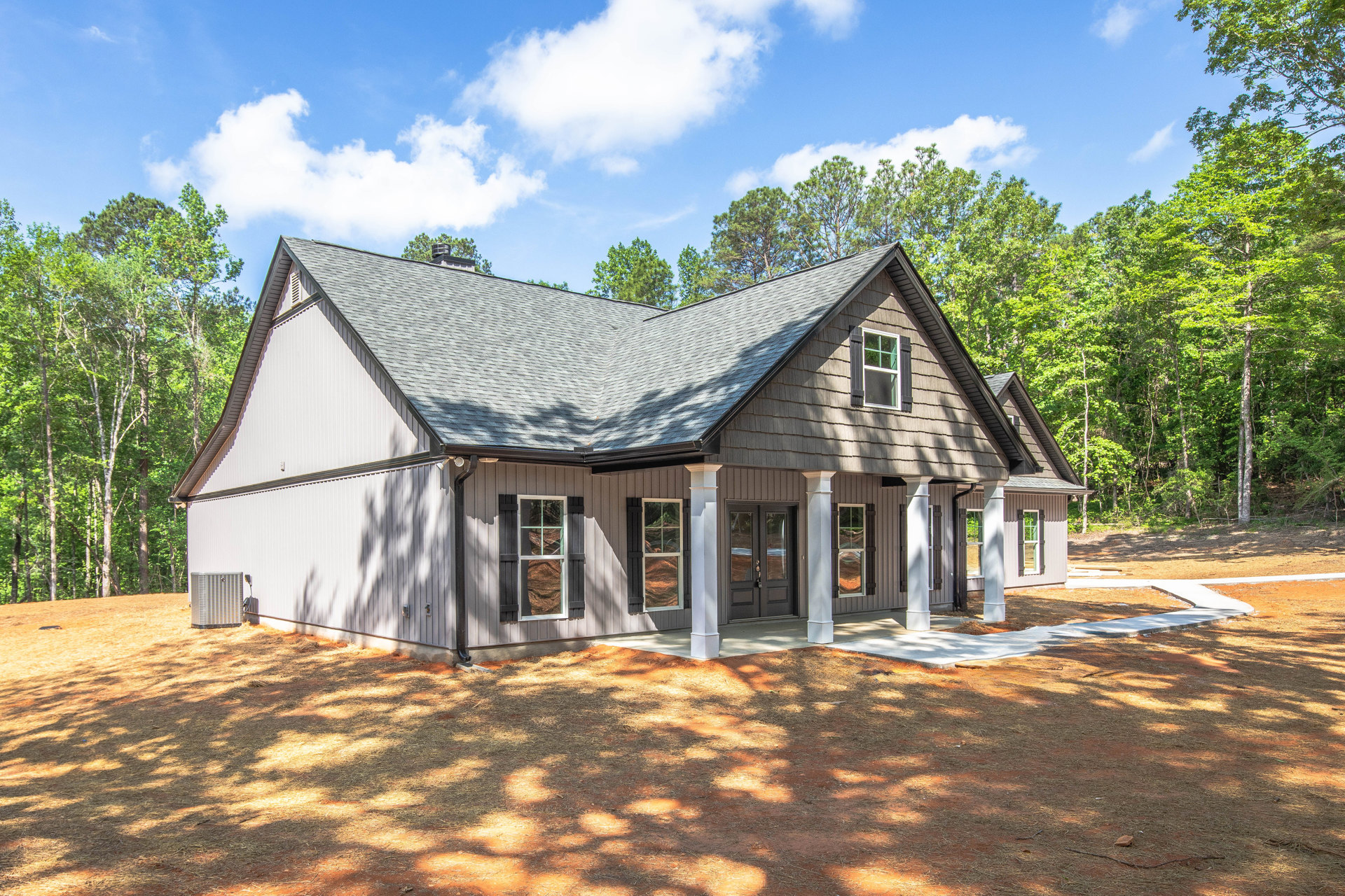 Two-story cottage with white columns, multiple windows, brown dirt landscaping, and mature trees in the background