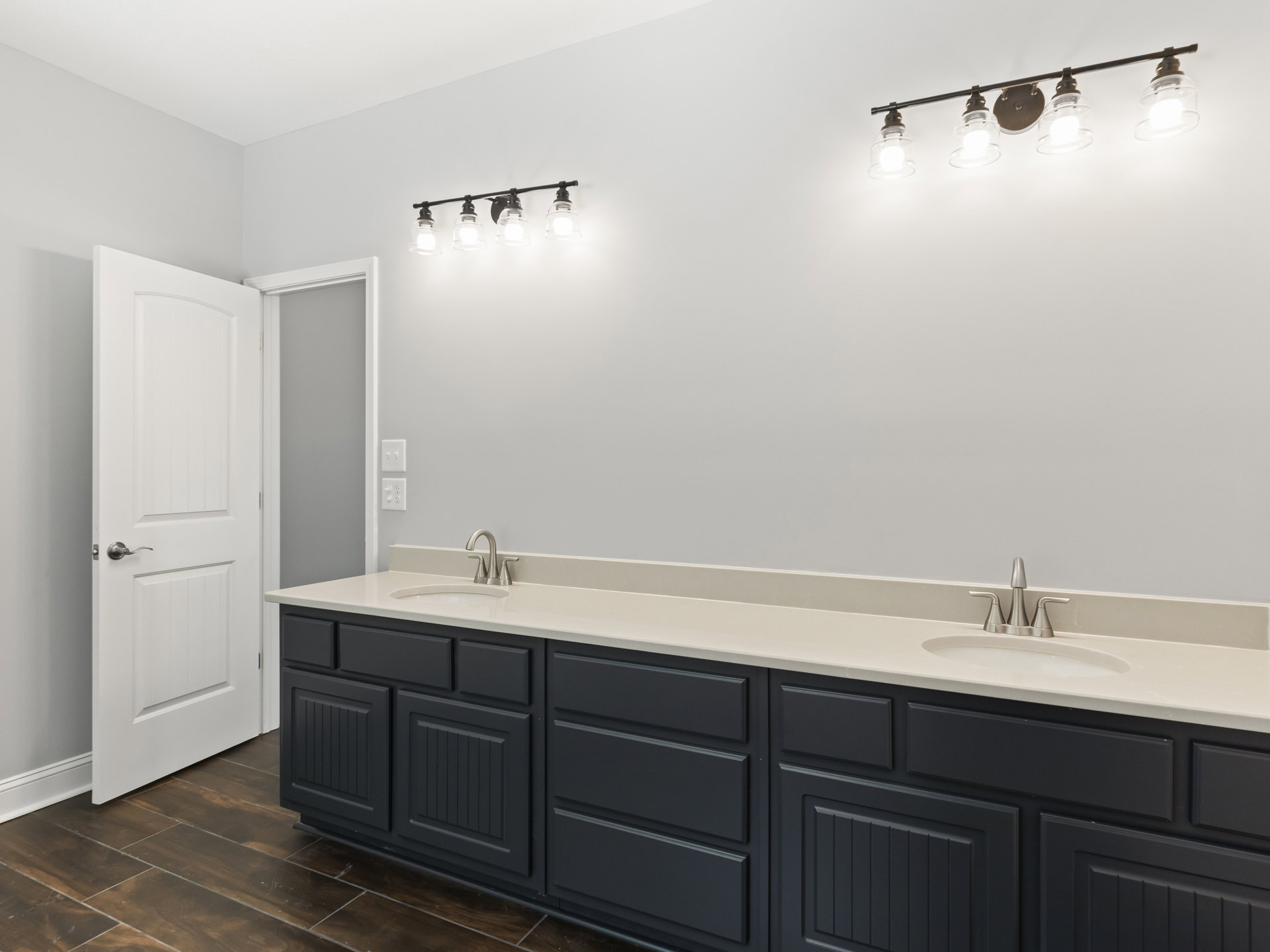 Bathroom with double sink vanity, wood flooring, white door with silver handle, three-light fixture above mirror, stone countertop with drawers, chrome faucet