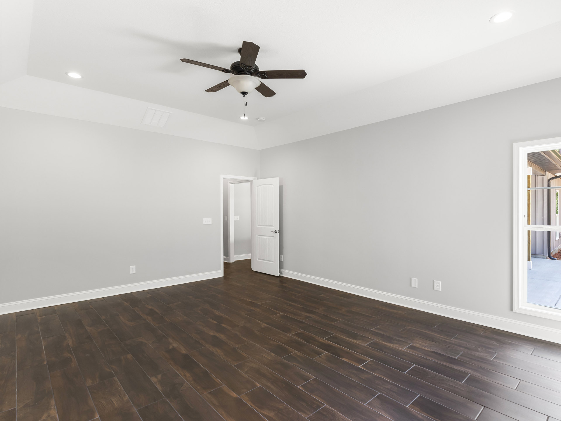 Ceiling fan with light fixture mounted above wood flooring, white walls, white door with handle, window with white frame, and light switch on wall