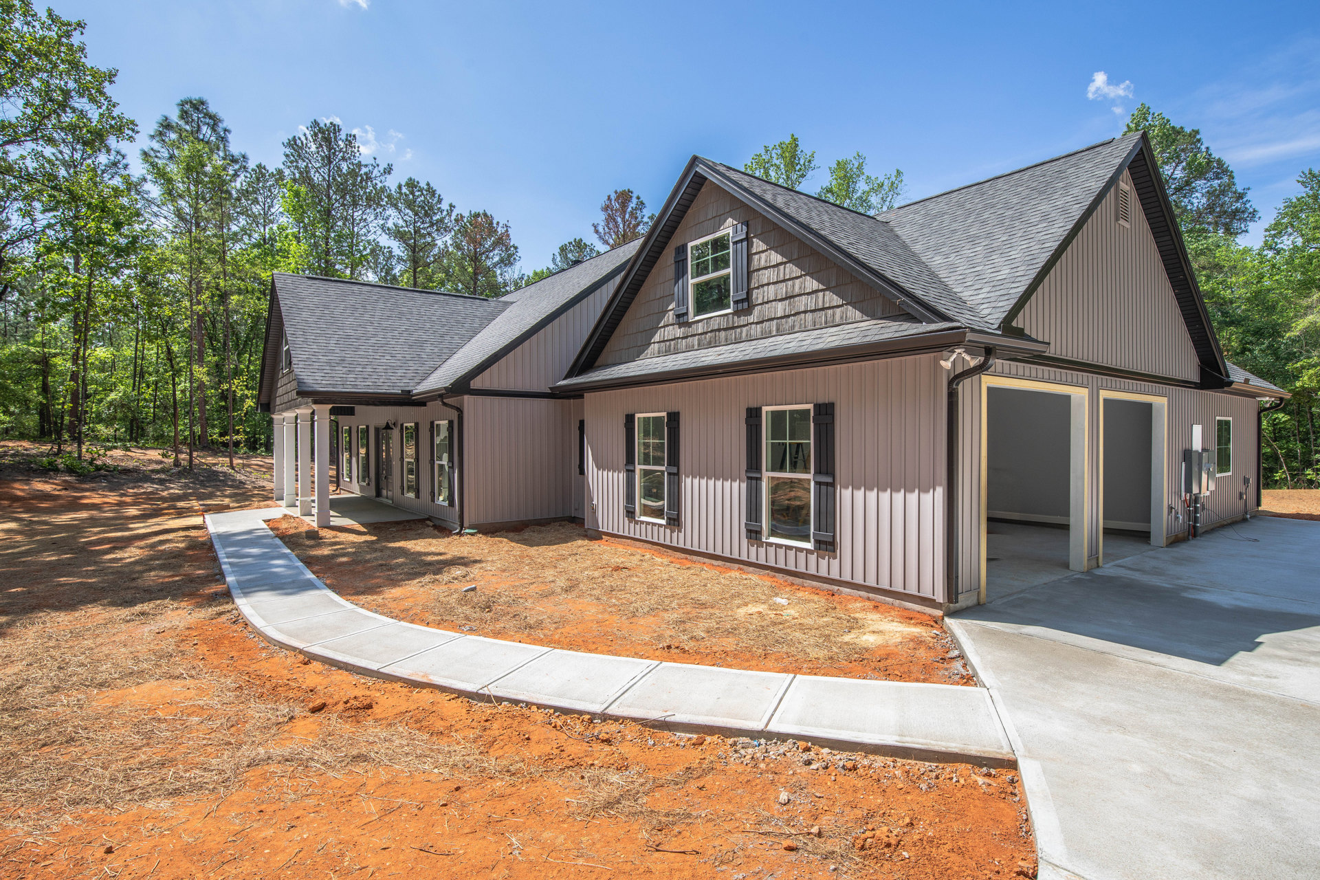 Two-story house with white-framed windows, attached garage, concrete driveway, and dirt sidewalk; mature trees and cloudy sky in background