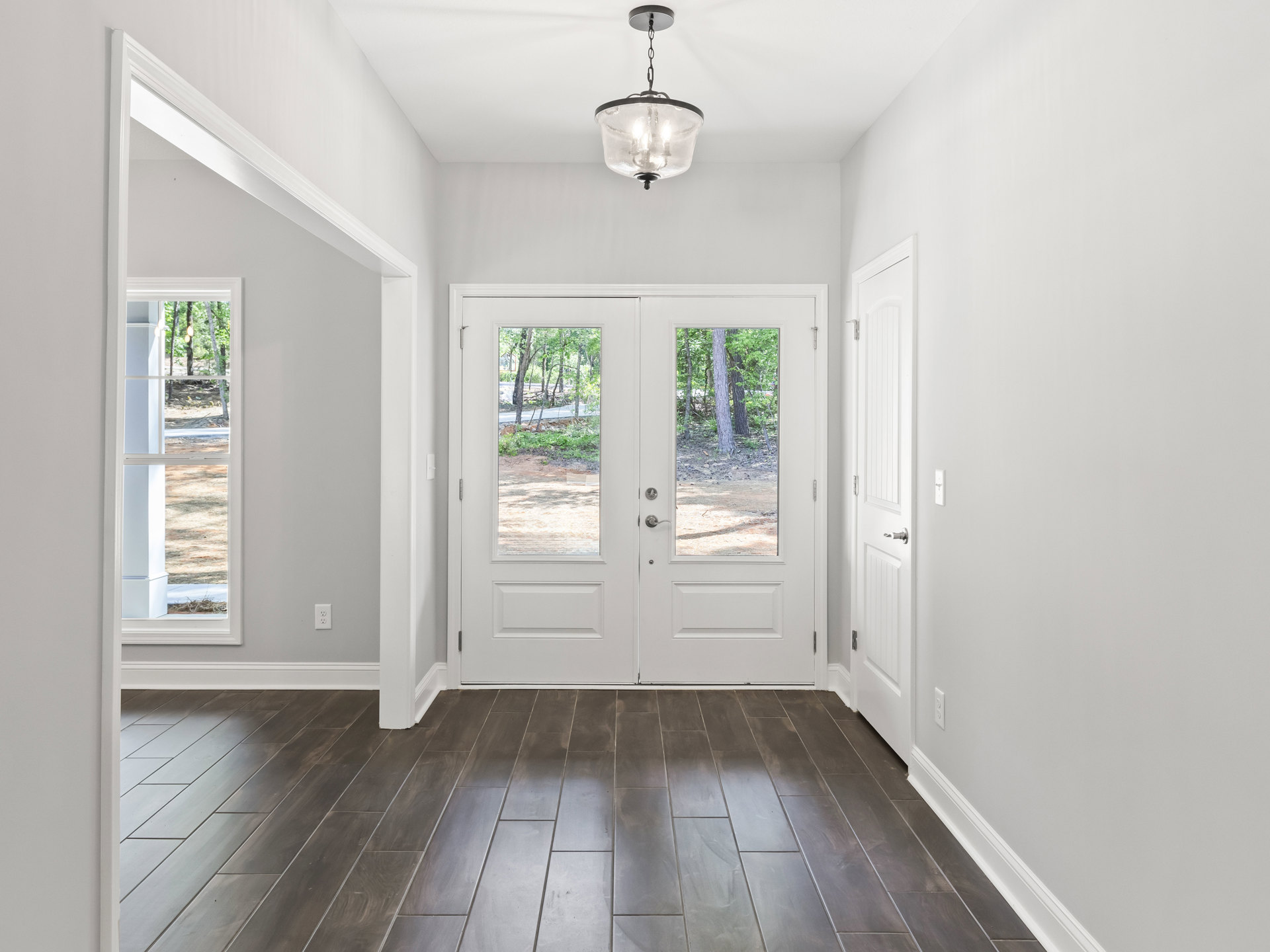 White hallway with wood floor, double glass-paneled doors, white walls, ceiling light fixture, and adjacent grey tile flooring.