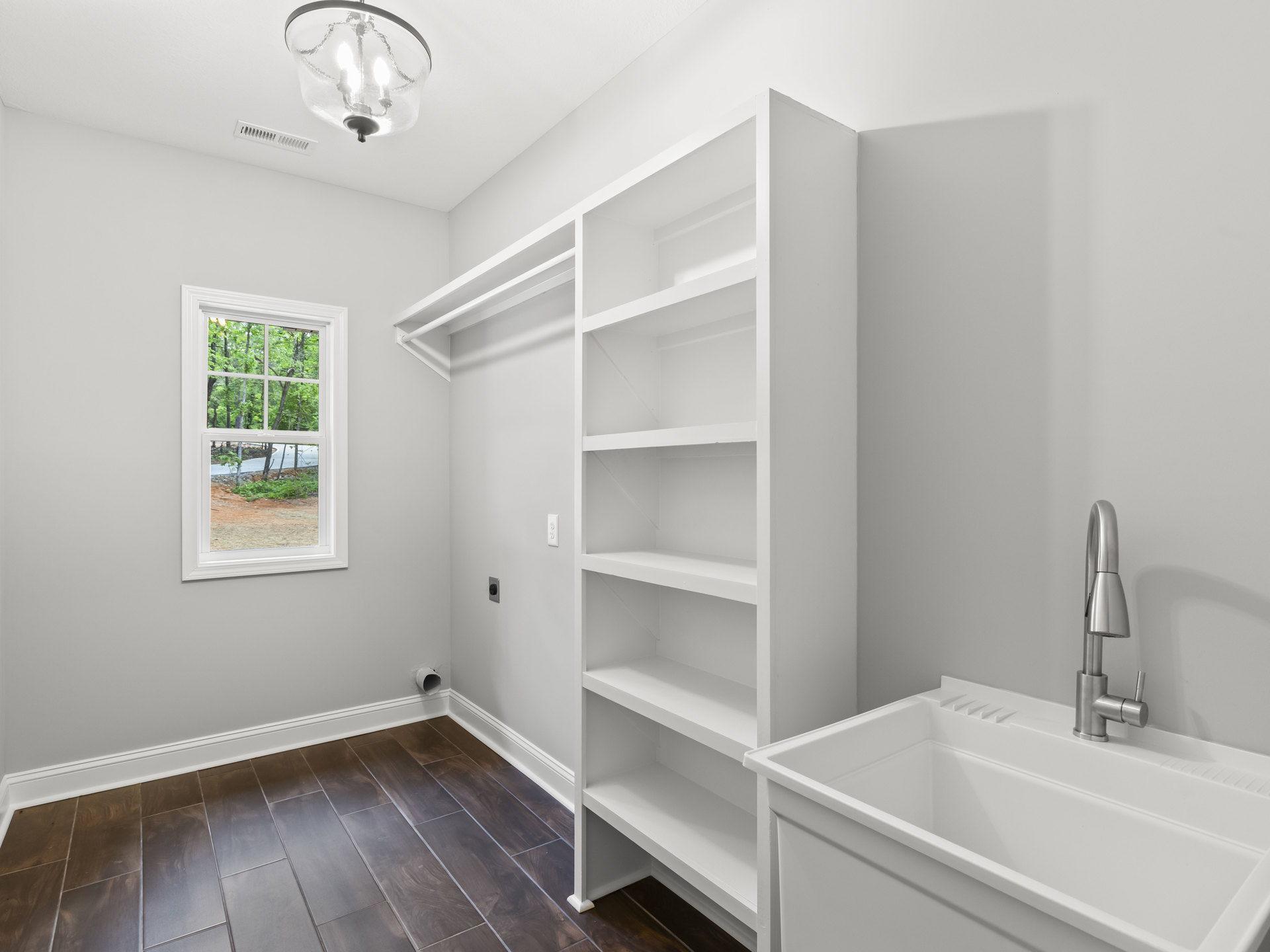 White bathroom with wood floor, white sink and silver faucet, open shelves, two-bulb light fixture, window showing trees, white trim along baseboard