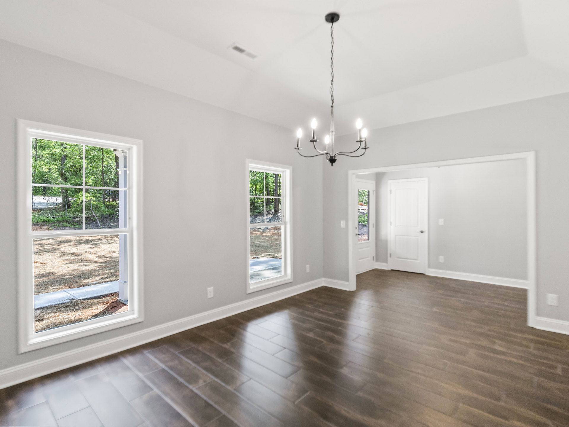 Hardwood floor room with white walls, large windows overlooking forest, silver-handled white door, ceiling-mounted chandelier with crystal accents