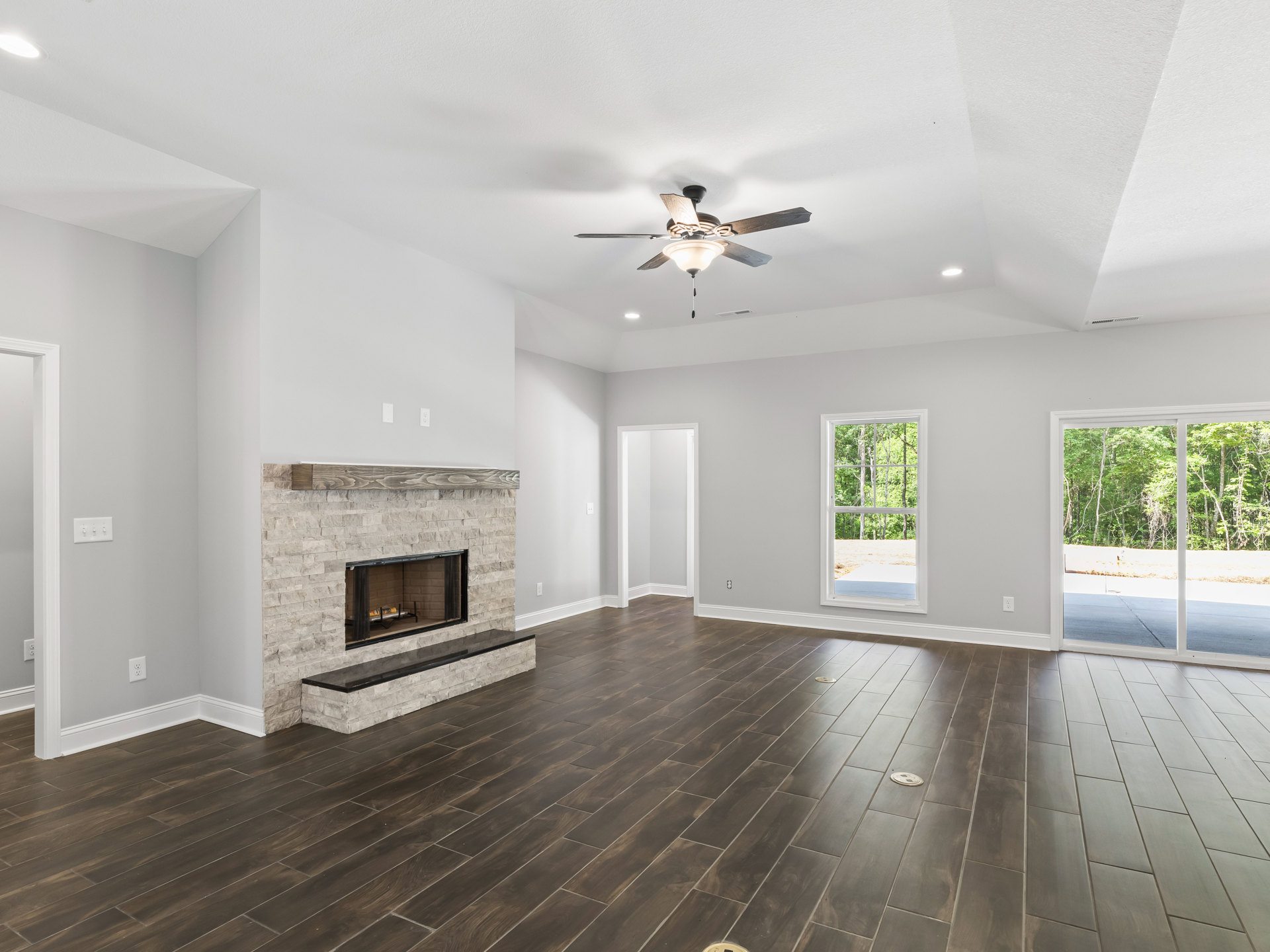 Living room with hardwood floors, central fireplace with black mesh screen, ceiling fan with light fixture, white door and window showing trees outside