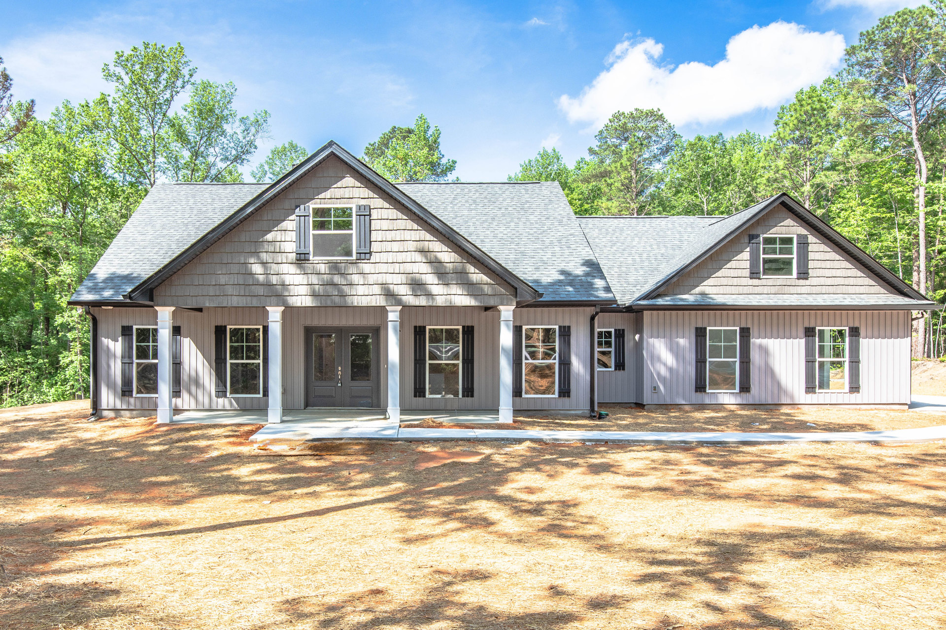 Wide front porch with white columns, large windows with white frames reflecting a road, light-colored siding, mature trees and blue sky in background