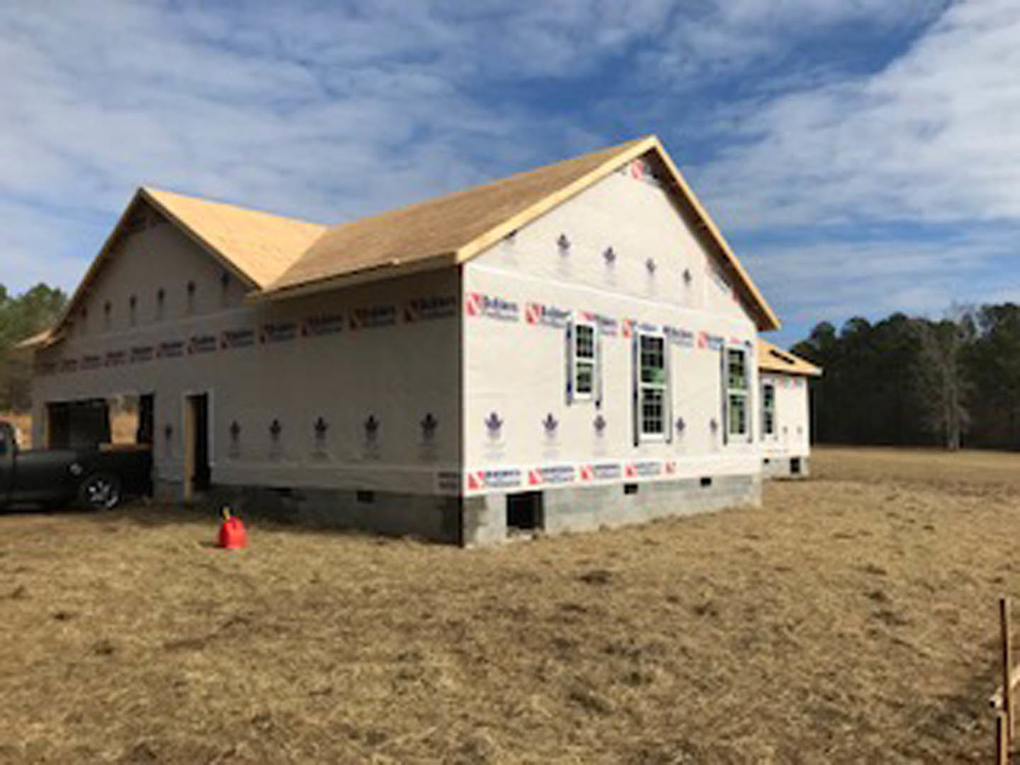 Framed house under construction with exposed plywood, red safety cone on dirt lot, dry grass, and trees in background under clear blue sky