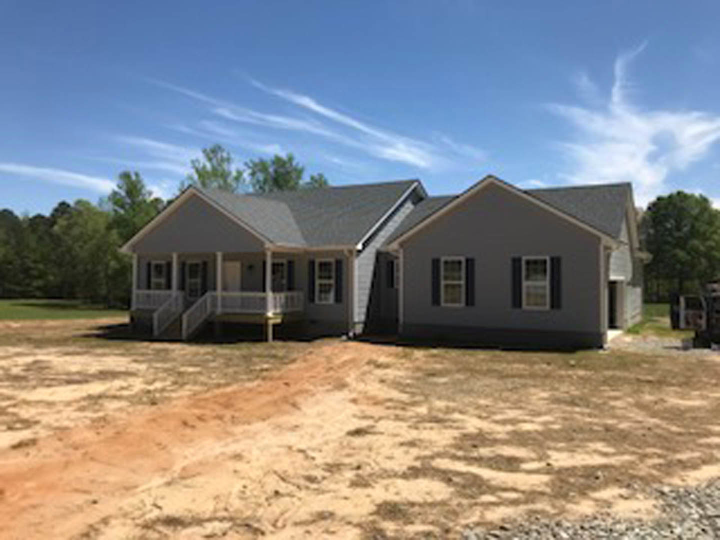 Modern home with open door, covered porch, dirt road leading to property, truck parked in distant field, blue sky with scattered clouds, person walking on balcony, gabled roof